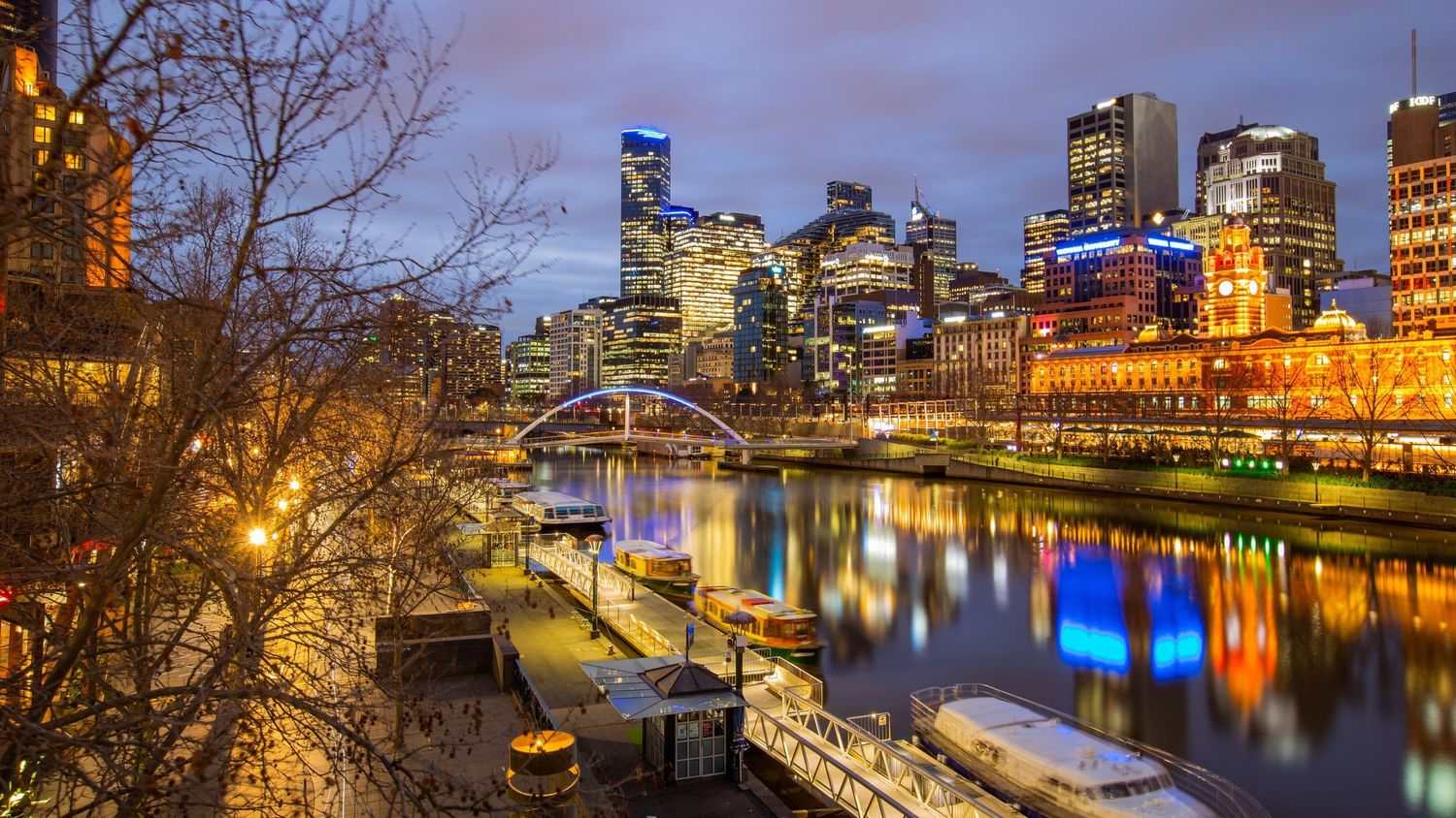 Panoramic cityscape of Melbourne skyline reflecting in Yarra River at twilight with illuminated skyscrapers and bridges.