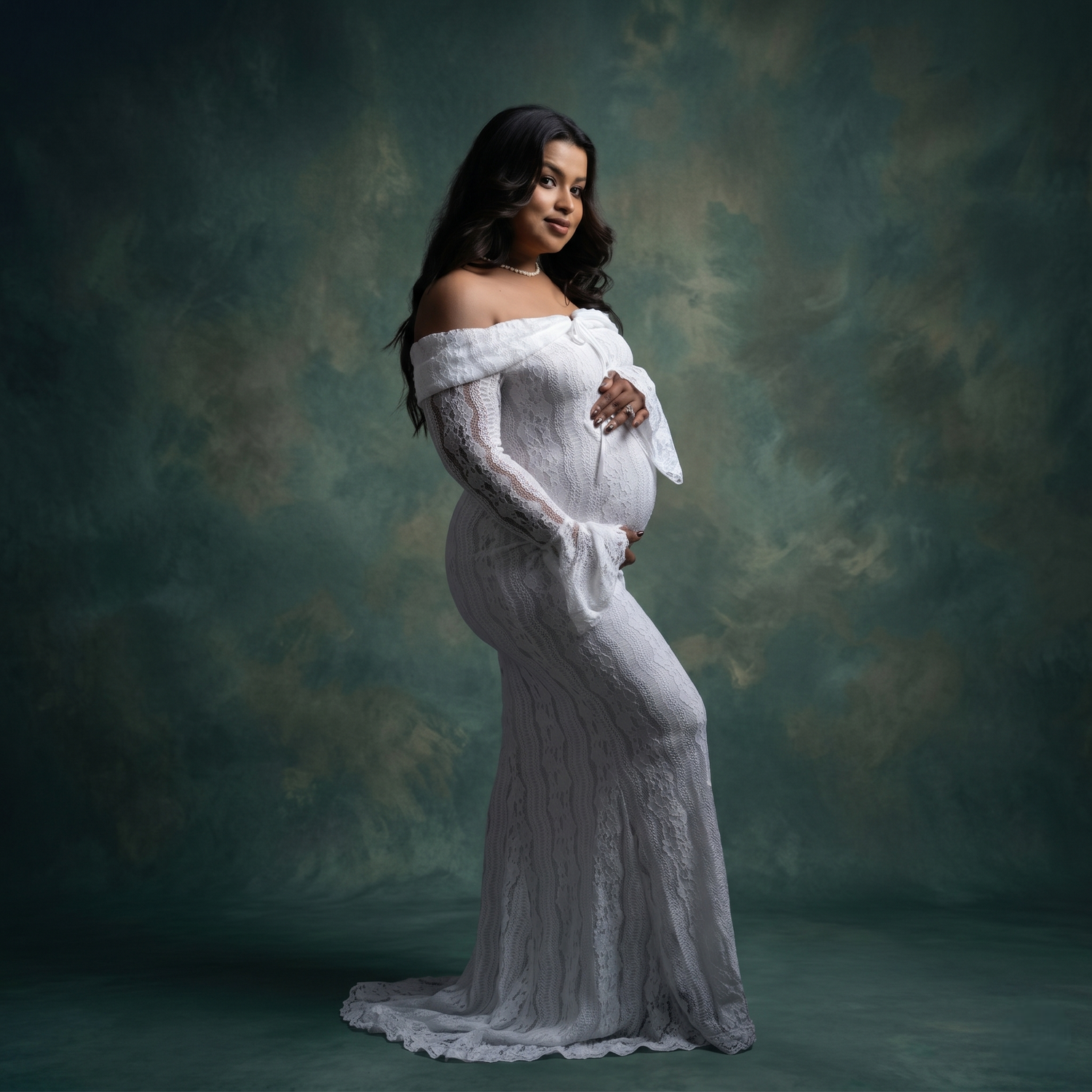 Pregnant woman in elegant white off-shoulder lace gown poses gracefully against a teal studio backdrop.