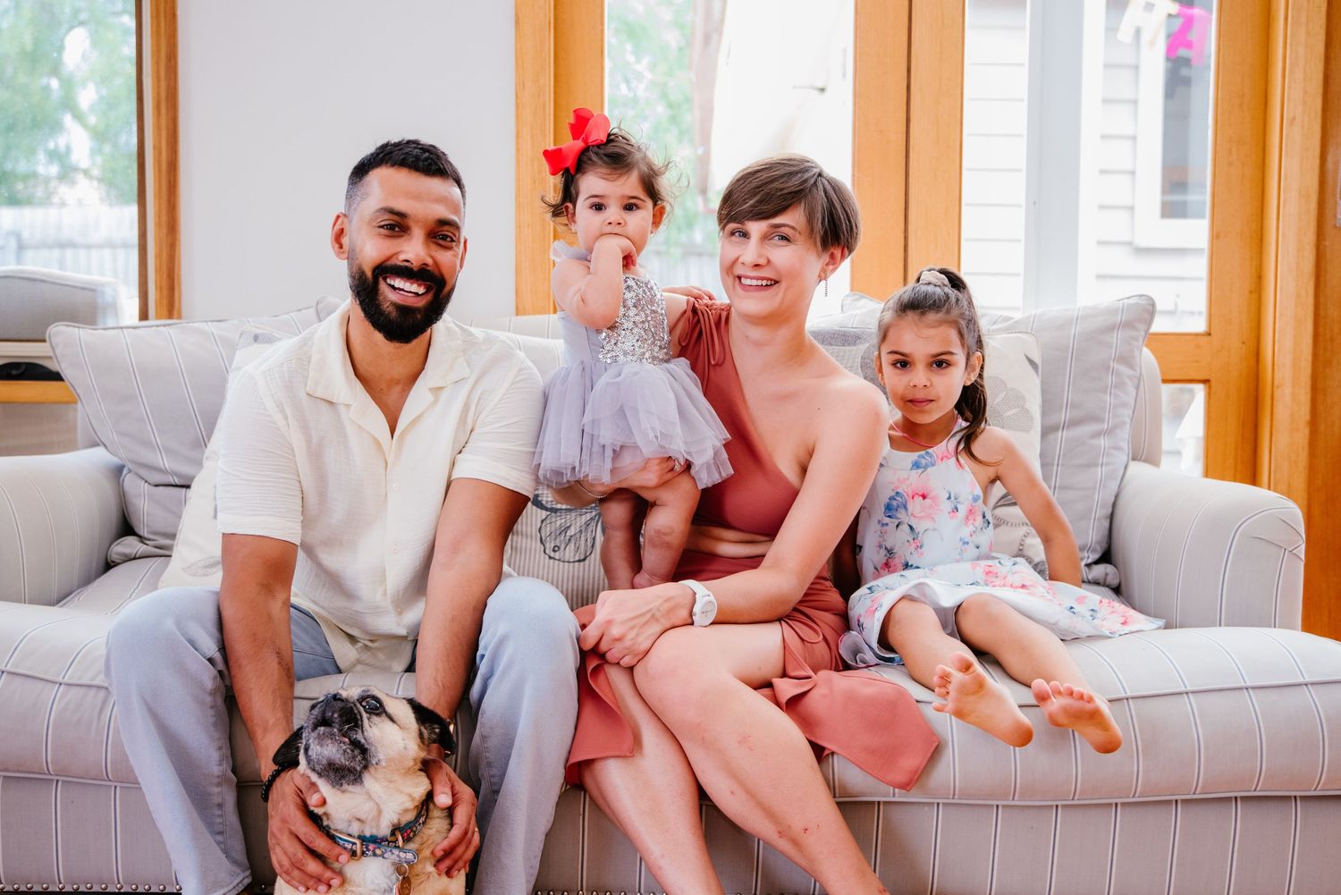 Family sits together on a grey couch in a sunlit living room with wooden window frames.