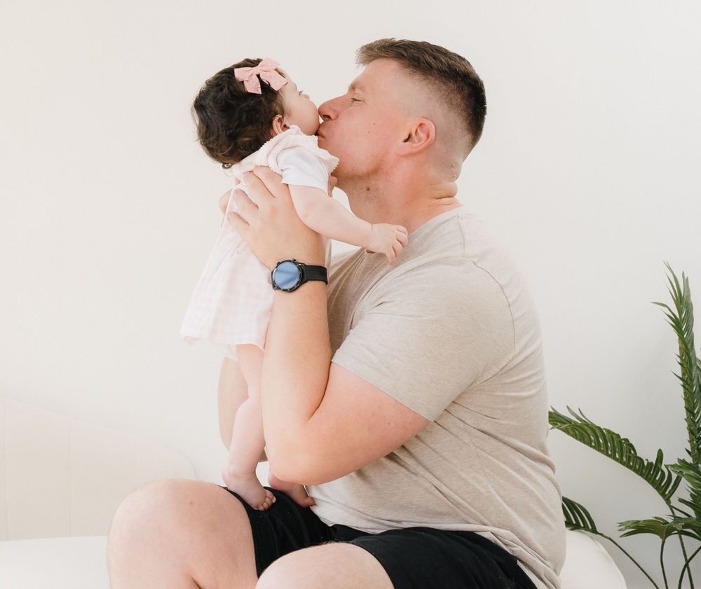 A loving father kisses his newborn baby while holding the infant up in a bright, minimalist white room.