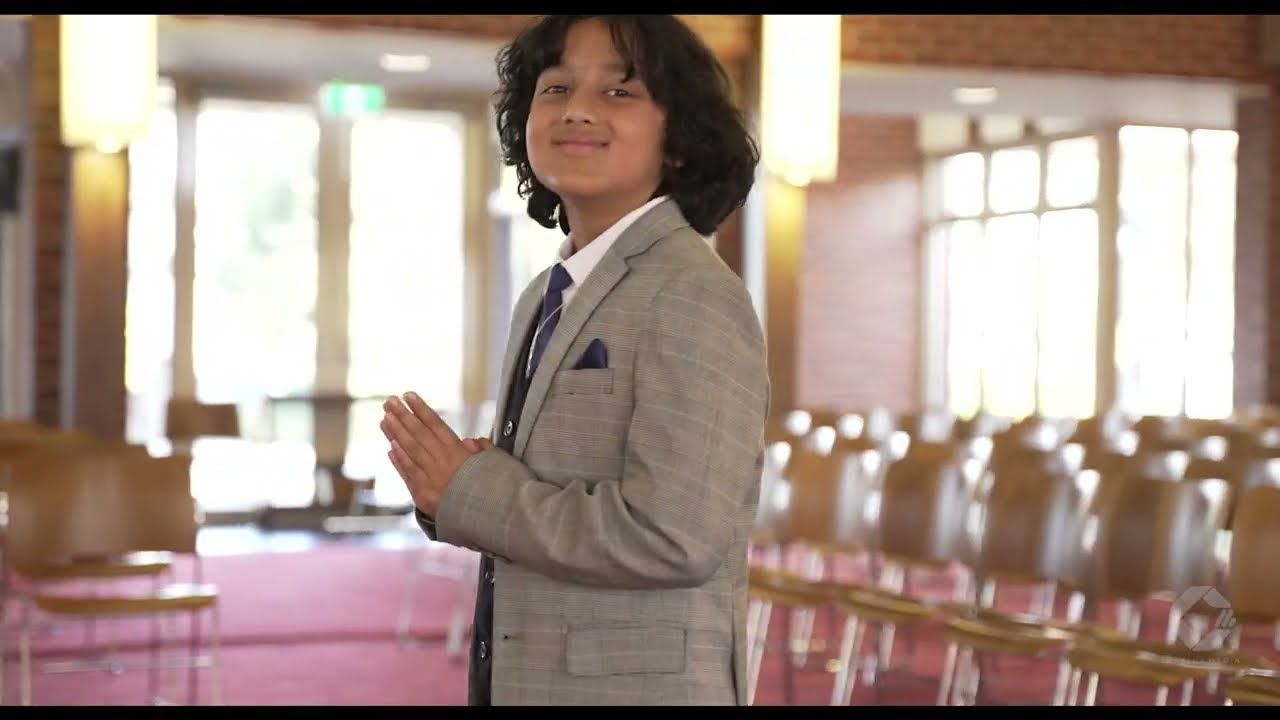 Young boy in gray suit smiling and clapping hands in an elegant hall with pink chairs.