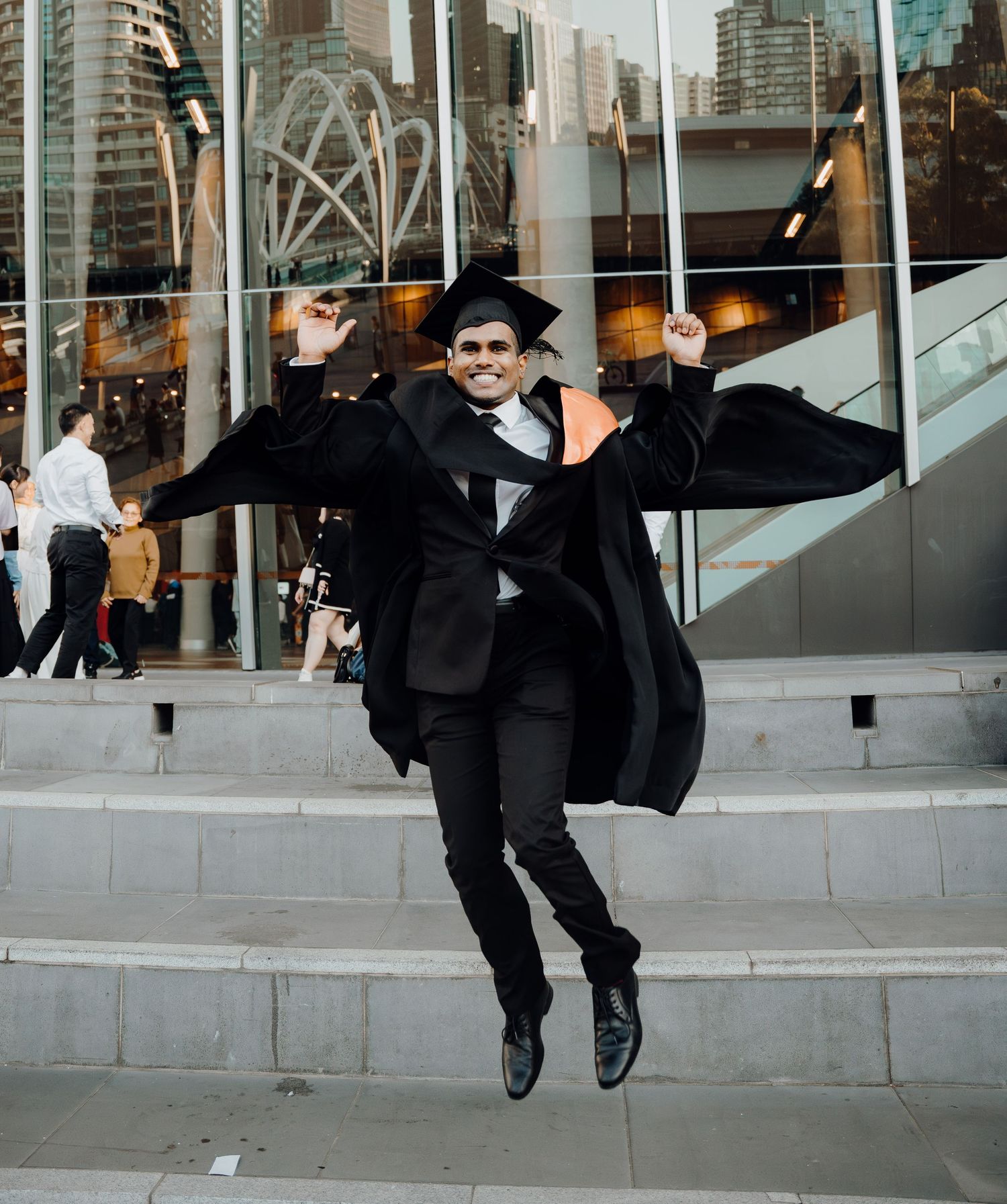 A person in a black suit and hat performs a jumping dance move on concrete steps in an urban setting.