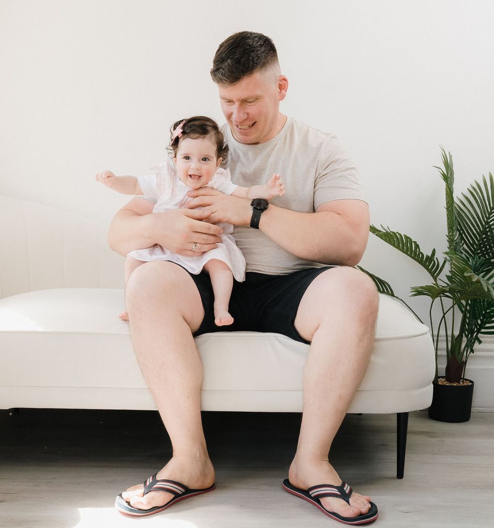 A smiling father sits on a white couch holding his happy baby girl in a bright, modern living room.