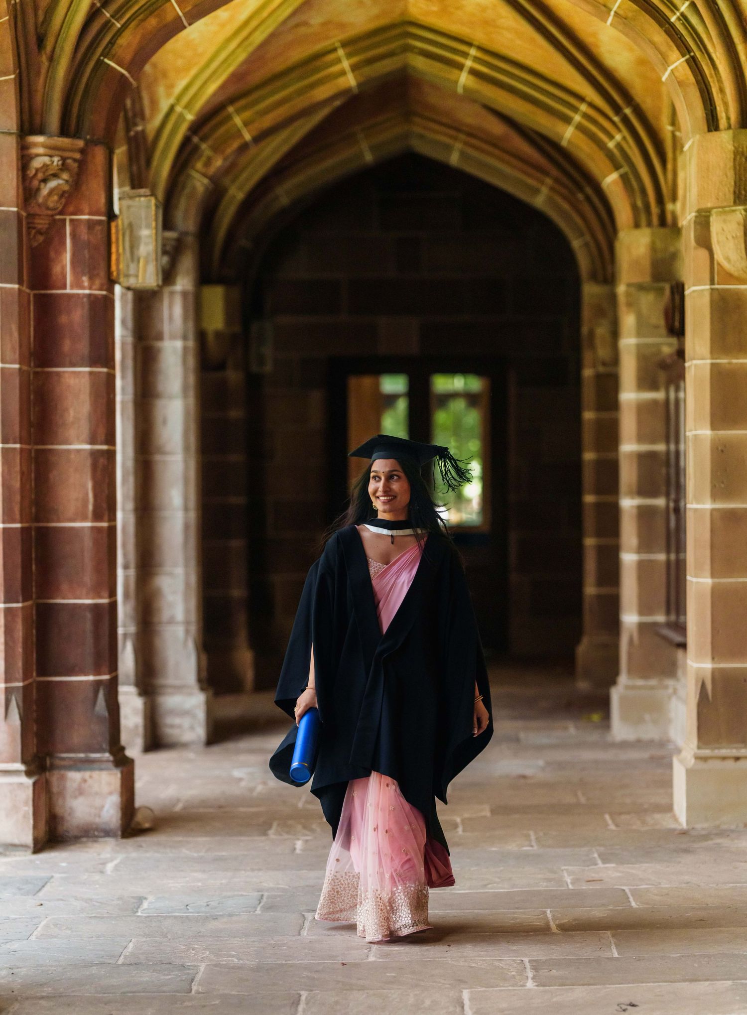 Graduate in traditional academic regalia stands in ornate stone archway of historic university building.