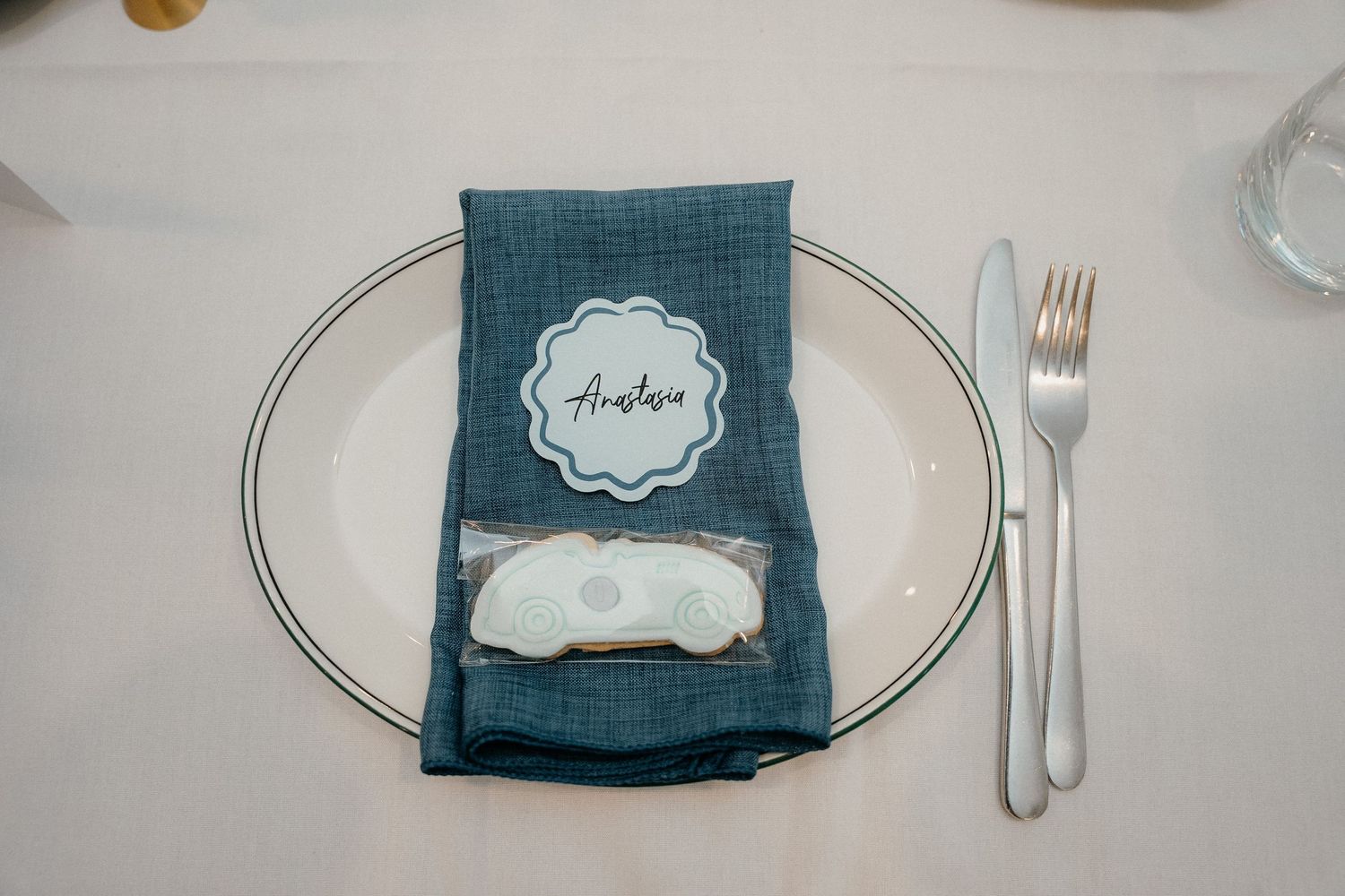 A teal blue napkin with a name card and place setting on a white plate with silverware.