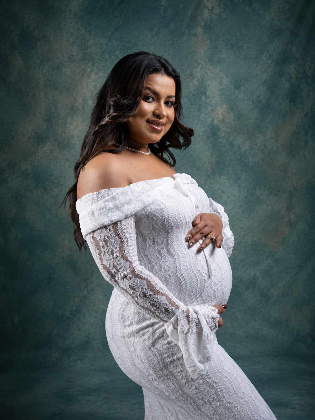 Pregnant woman in white off-shoulder lace gown posing for maternity portrait against dark teal studio backdrop.