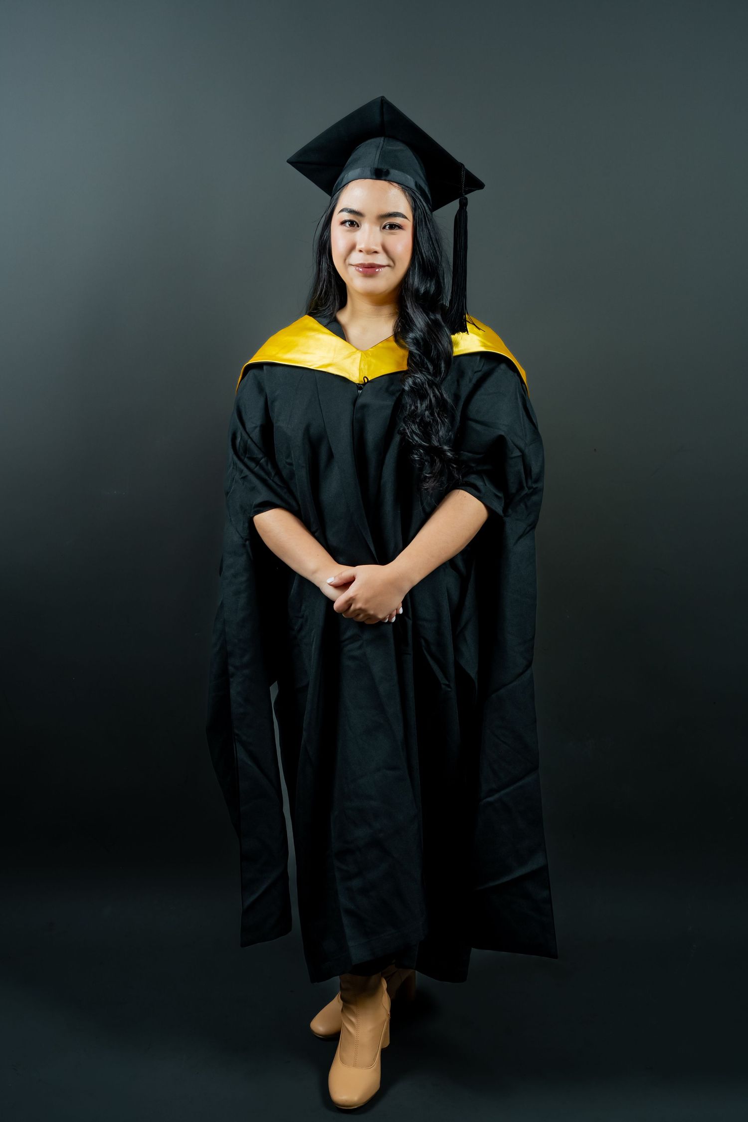 Graduate wearing black cap and gown with yellow sash poses against dark background for professional portraits.