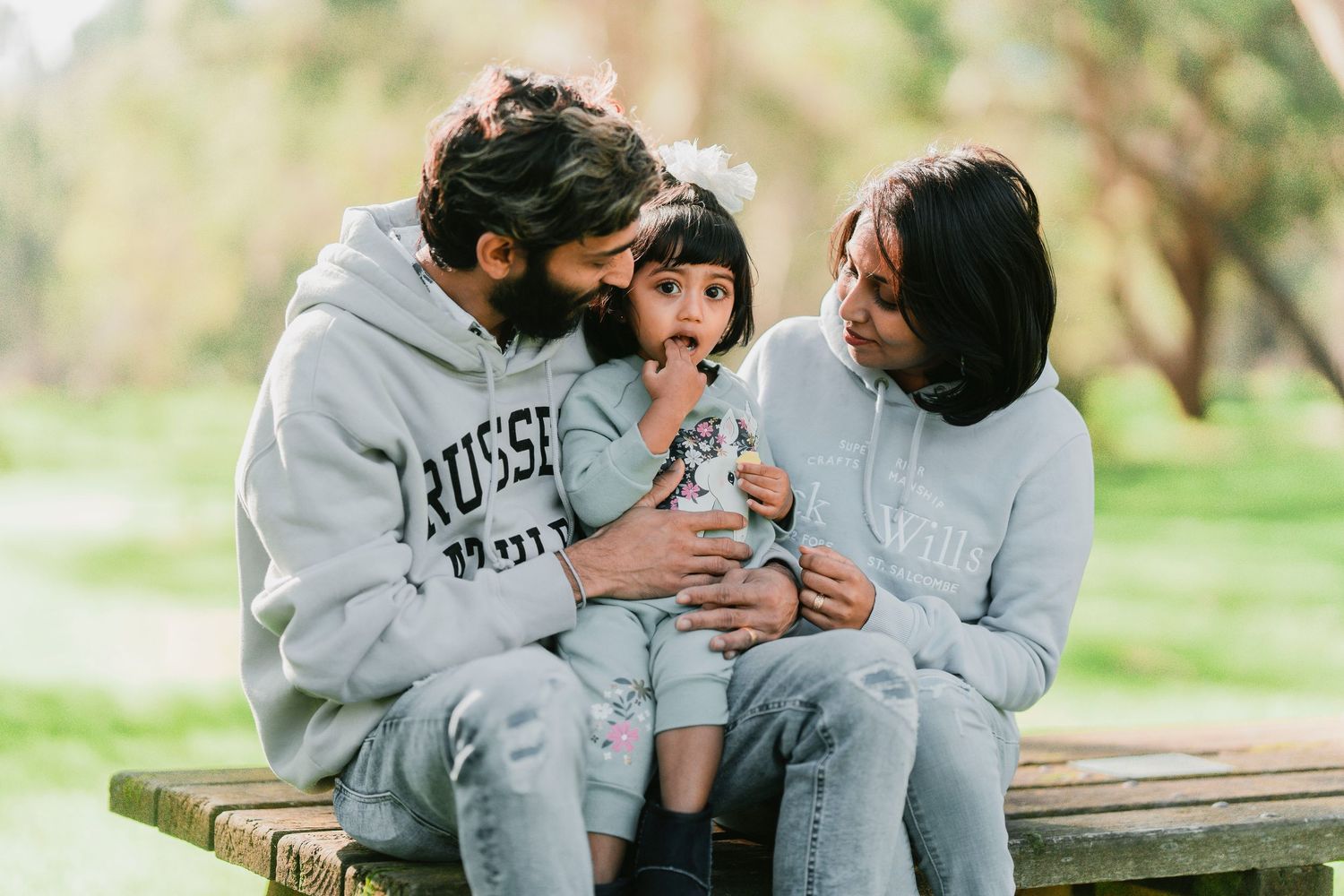 A family wearing matching gray hooded sweatshirts sits together on a wooden bench in a park setting.