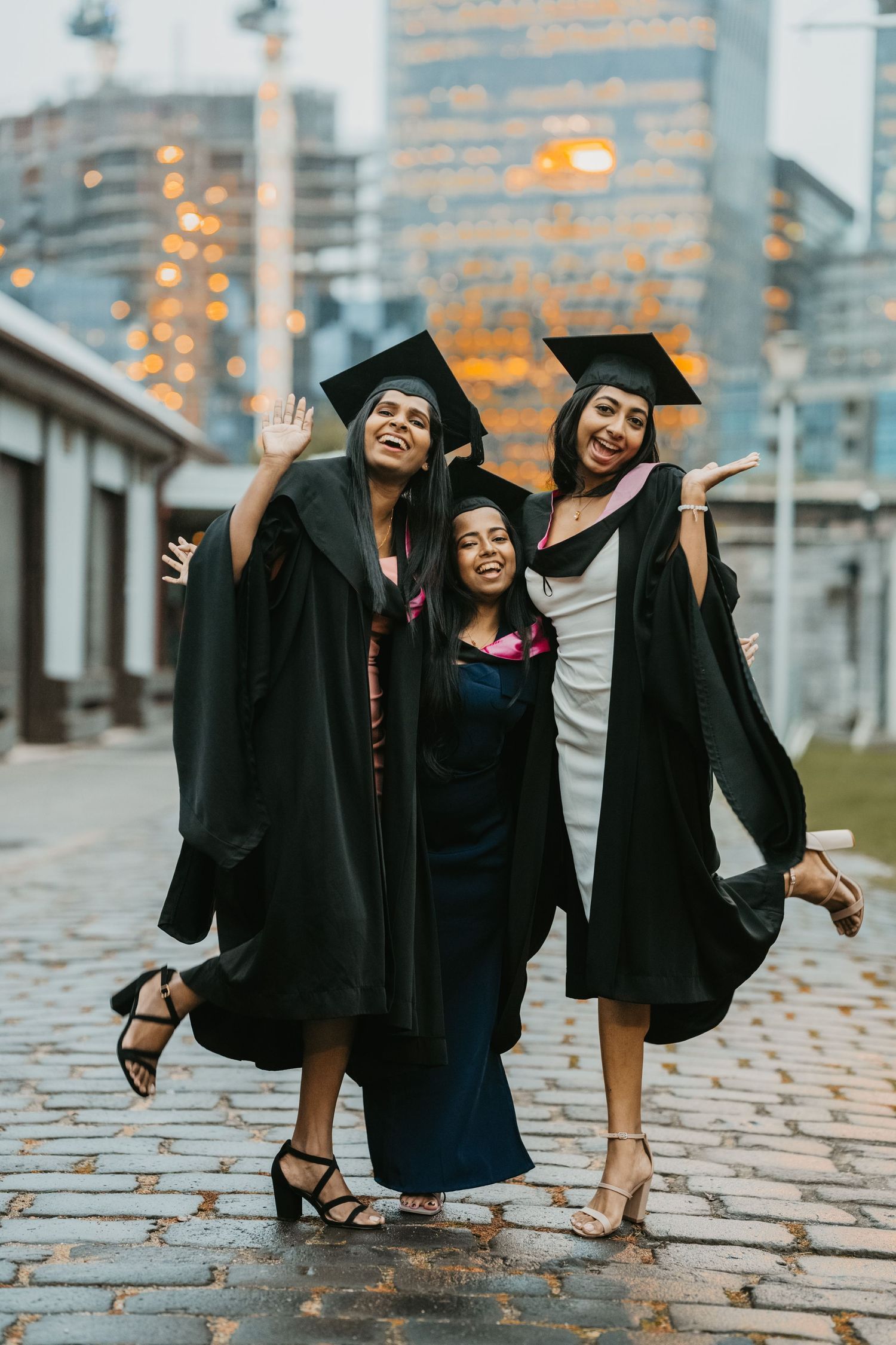 Three women in graduation gowns and caps celebrate joyfully on a cobblestone street with city buildings in the background.