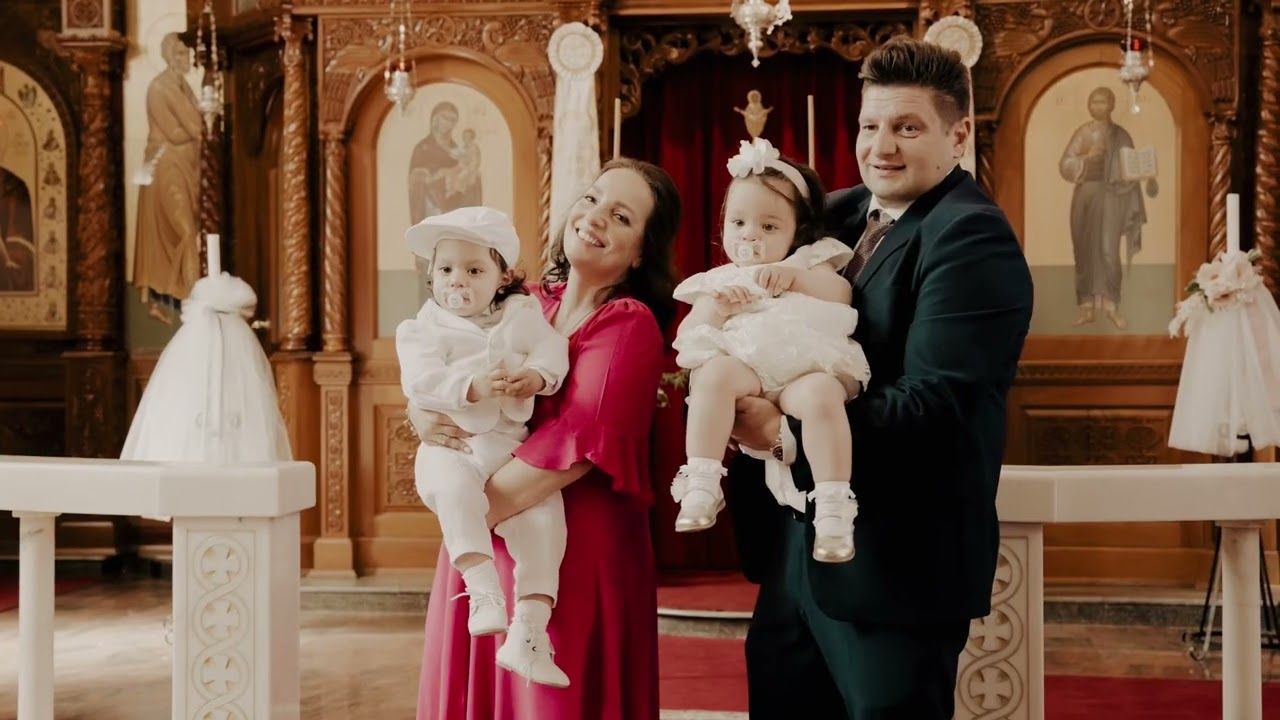 Family posing in Orthodox church during baptism ceremony, parents holding two babies dressed in white christening outfits.