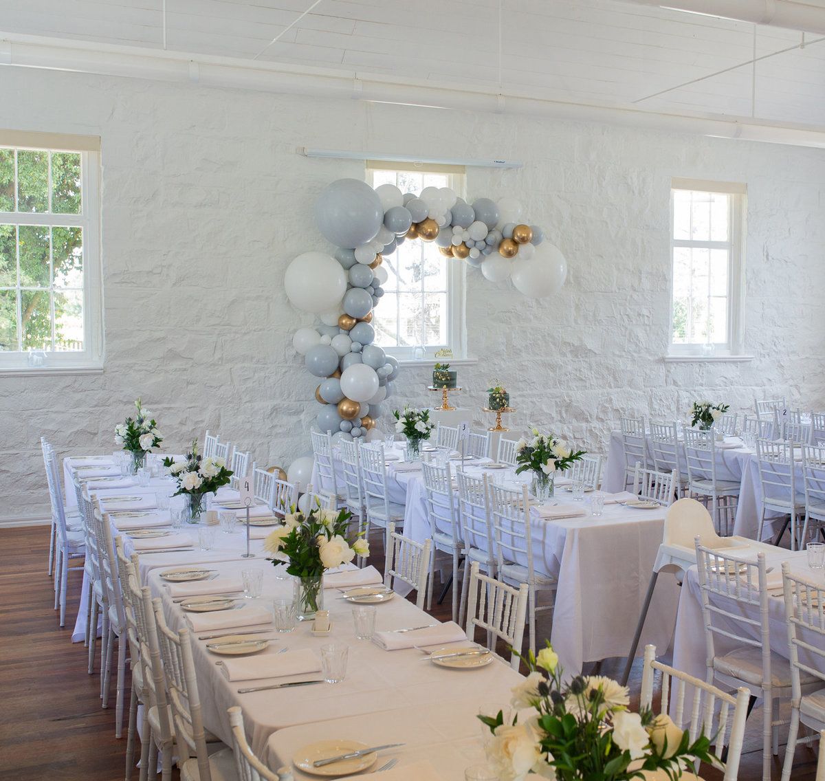 White wedding reception venue with long tables set with white linens and floral centerpieces in a bright airy room.