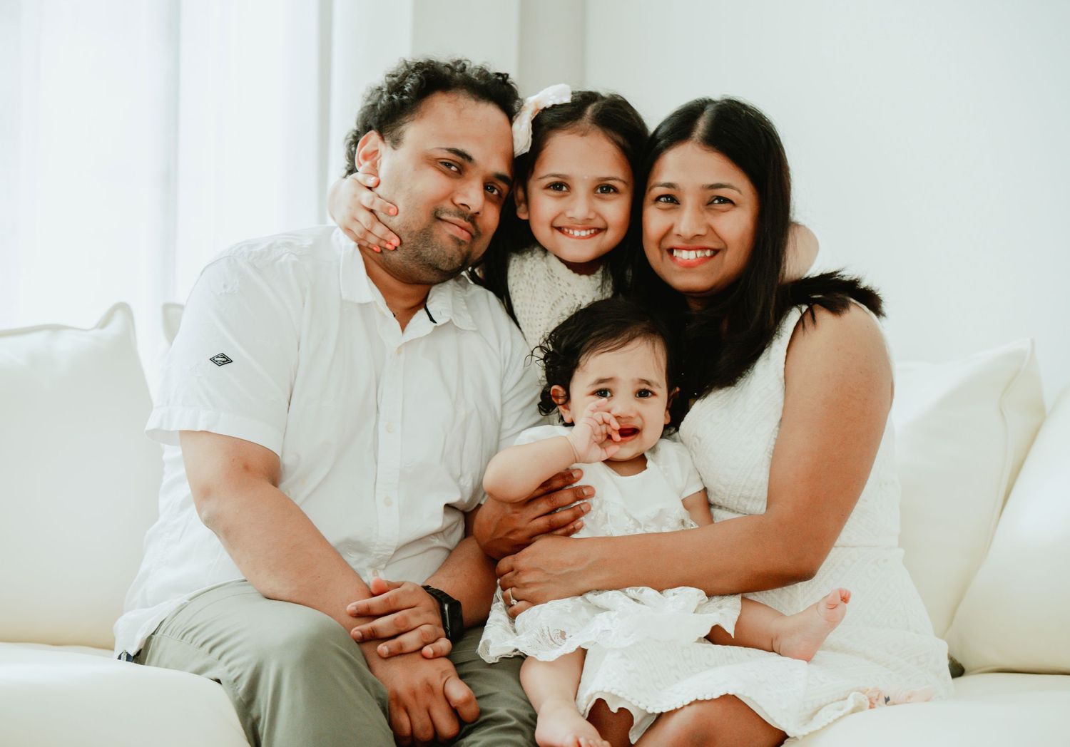 A happy family of four sits together on a white couch, smiling warmly at the camera.
