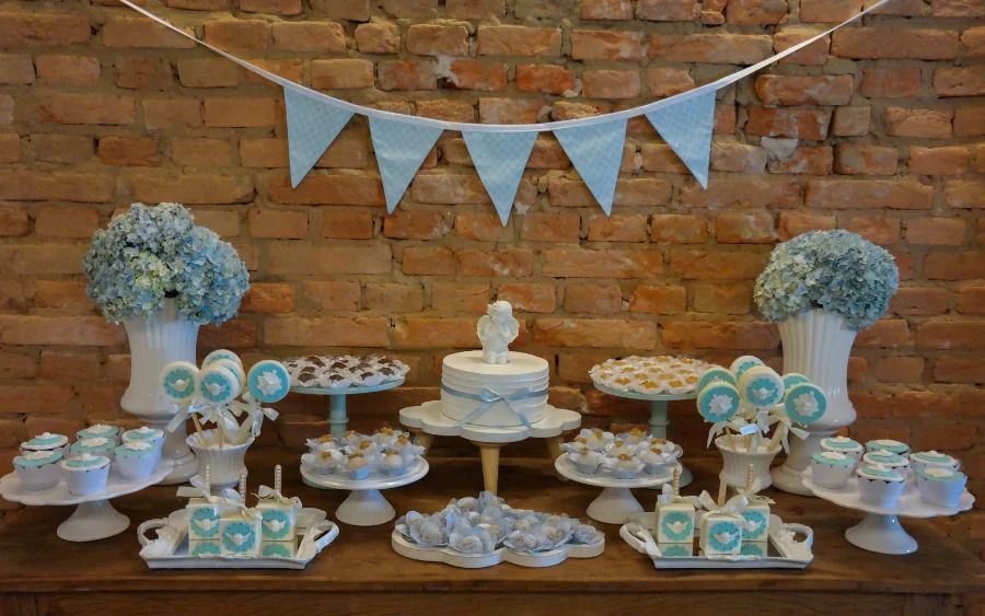Elegant dessert table with blue and white decorations featuring a cake, cupcakes, and floral arrangements against brick wall.