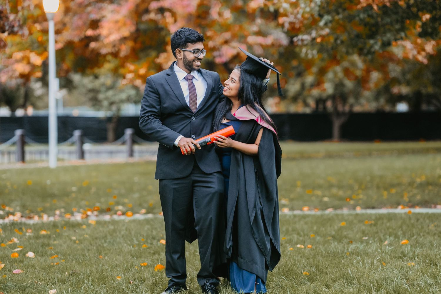A couple in formal attire walks together on a grassy lawn during autumn with fallen leaves in the background.