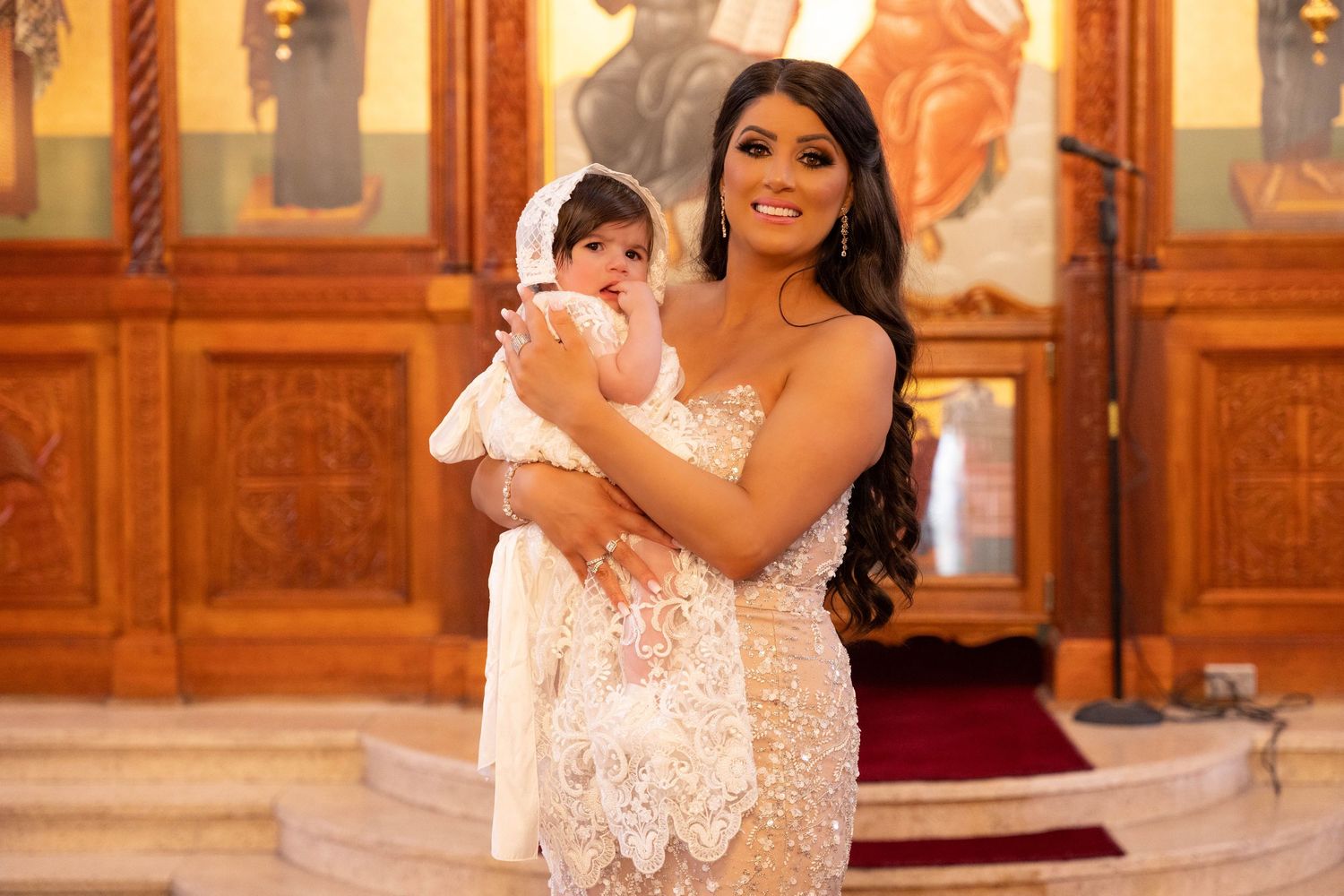 A mother holding a baby in white christening attire stands inside an ornate church with wooden paneled walls and lighting.