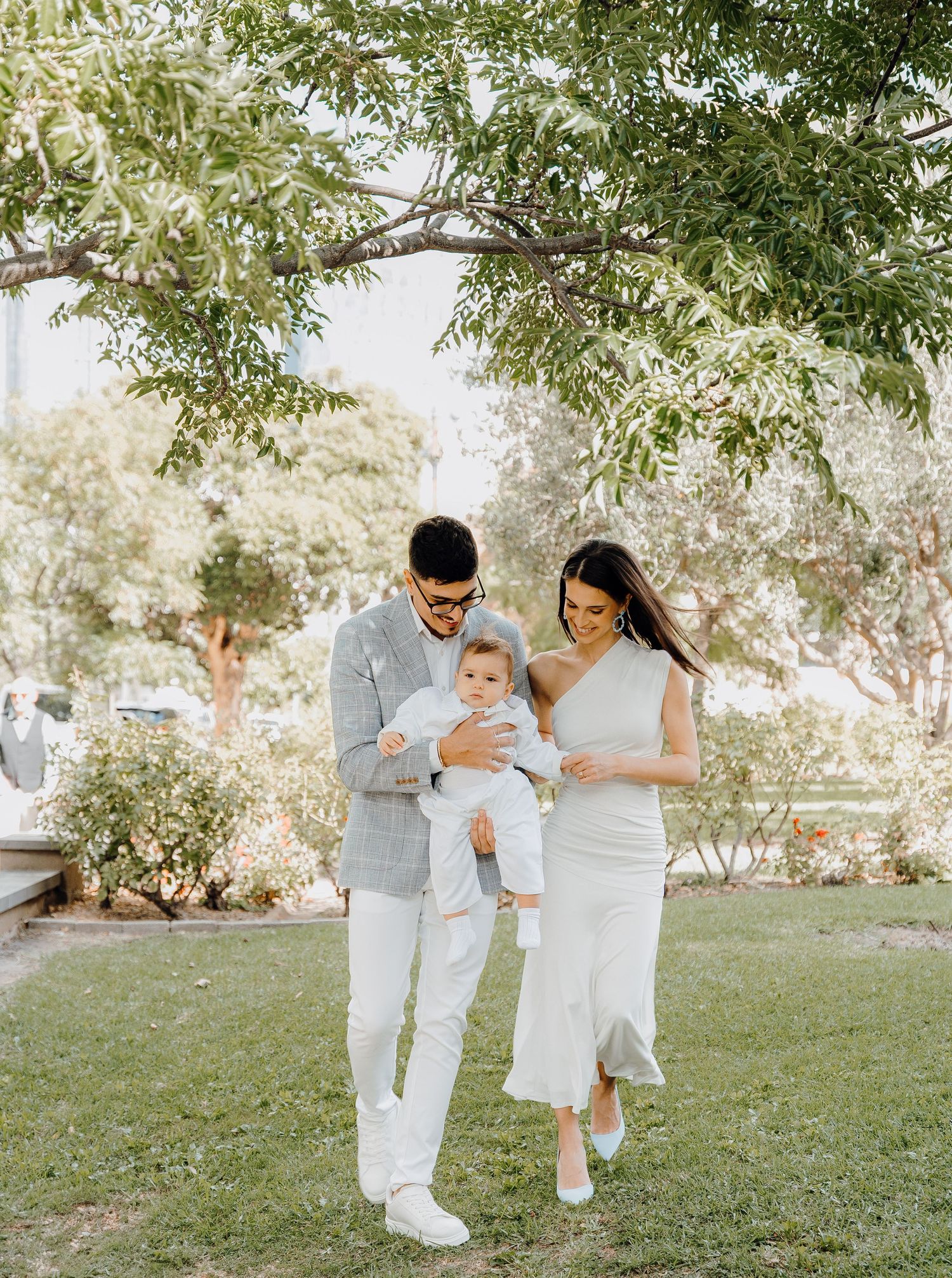 A family in white outfits plays together on grassy lawn under tree branches during an outdoor photo session.
