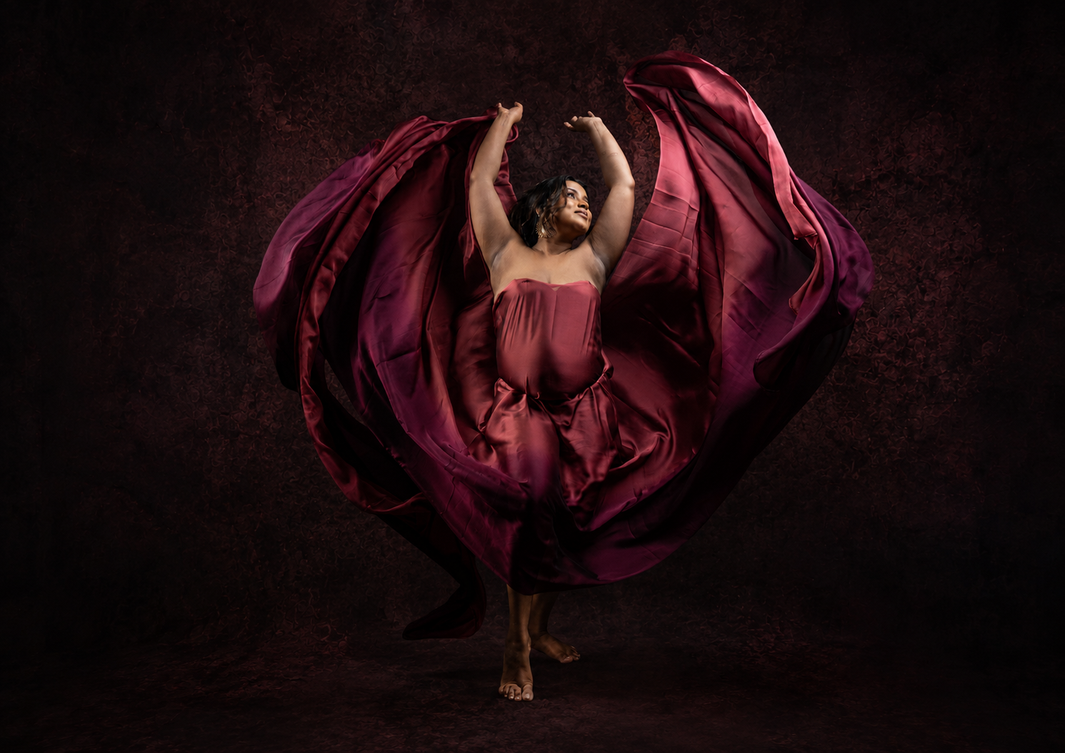 A dancer in a flowing burgundy gown with fabric billowing like wings against a dark dramatic background.