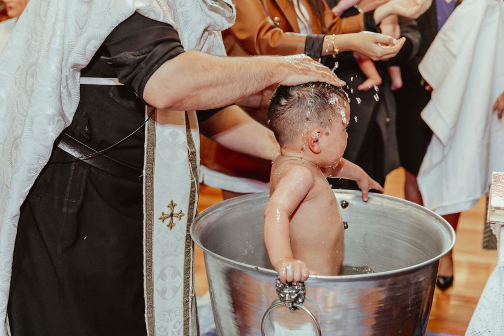 Priest performing Orthodox Christian infant baptism, pouring water over baby in traditional metal baptismal font.