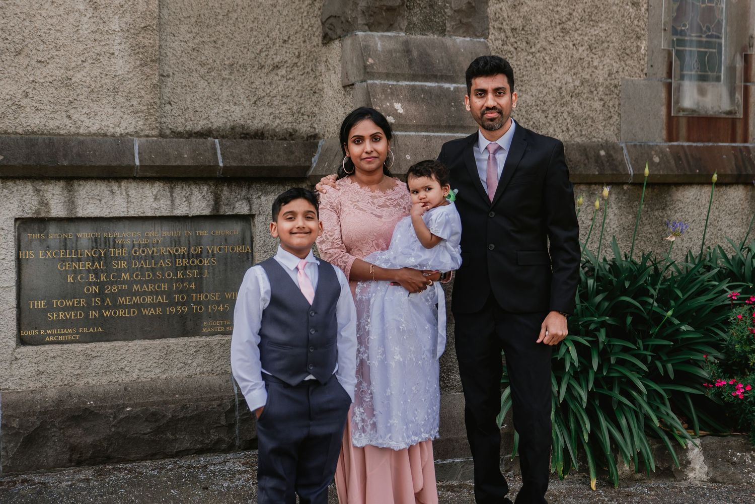 A group poses together at a formal event in front of a stone wall with green plants.