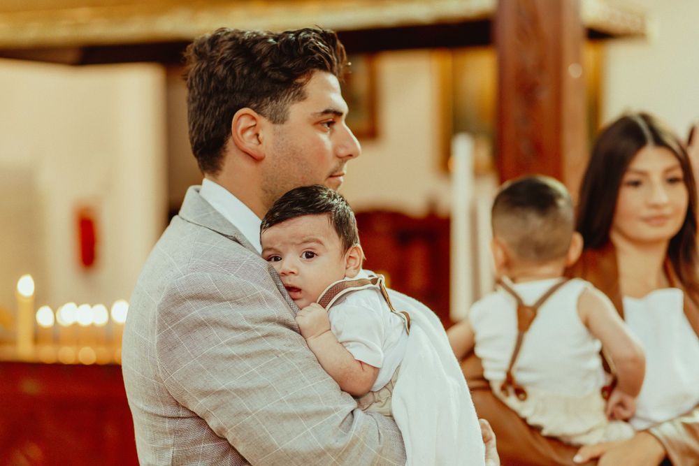 Man in gray suit holds baby at church baptism ceremony, woman and child seated in background.