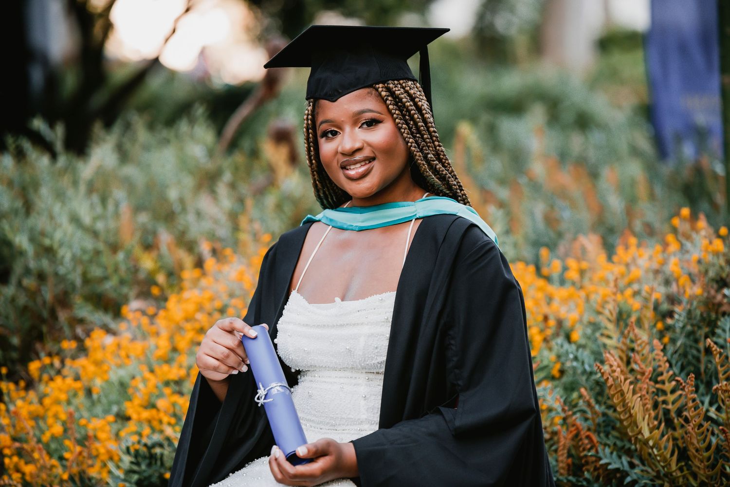 Graduate in cap and gown poses with diploma among vibrant yellow flowers and greenery in an outdoor garden setting.