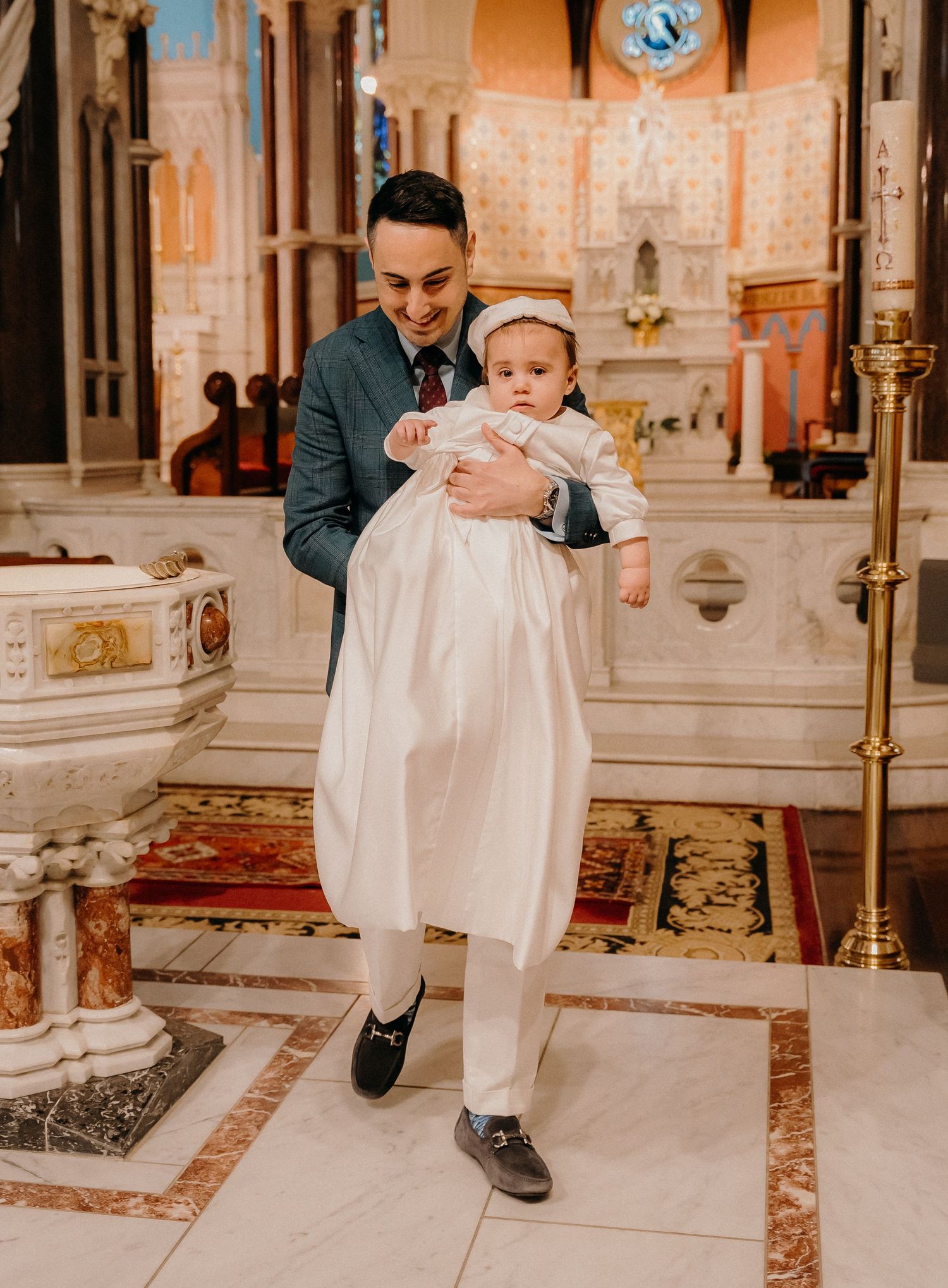 Baby in white christening gown crawls on ornate church floor during baptism ceremony.