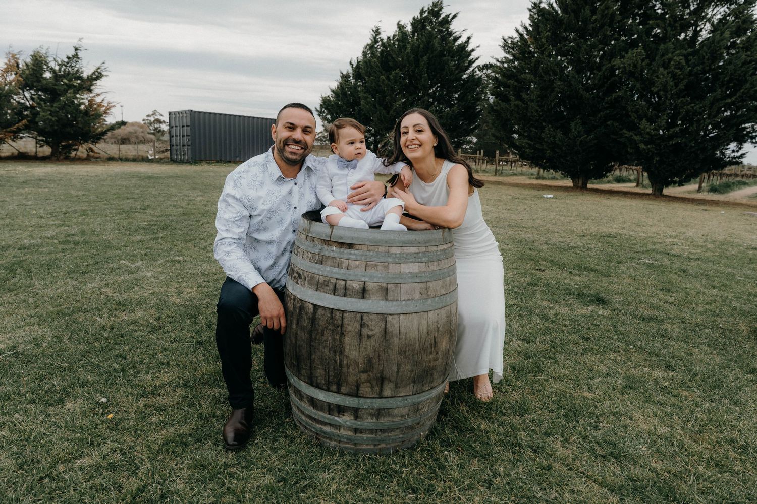 A couple poses playfully around a rustic wine barrel in a grassy outdoor setting at sunset.