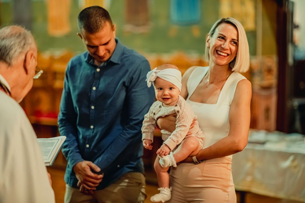 Young family with baby at a baptism ceremony, smiling mother holding infant in white headband with father beside her.