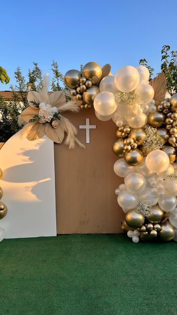 Elegant balloon arch with white and gold ornaments decorates a brown backdrop with a cross for a religious celebration.