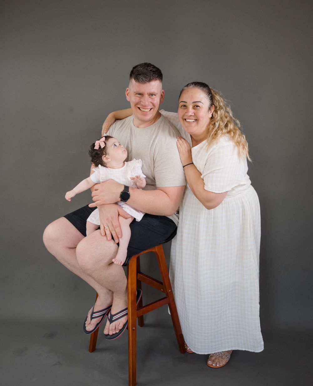 A smiling couple sits on a stool against a gray backdrop, lovingly holding their newborn baby dressed in white.