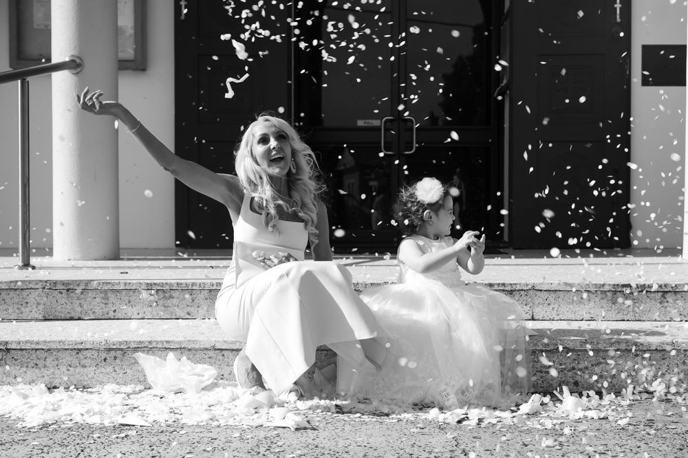 Two women in white dresses laugh joyfully as confetti showers around them on steps in a black and white wedding photo.