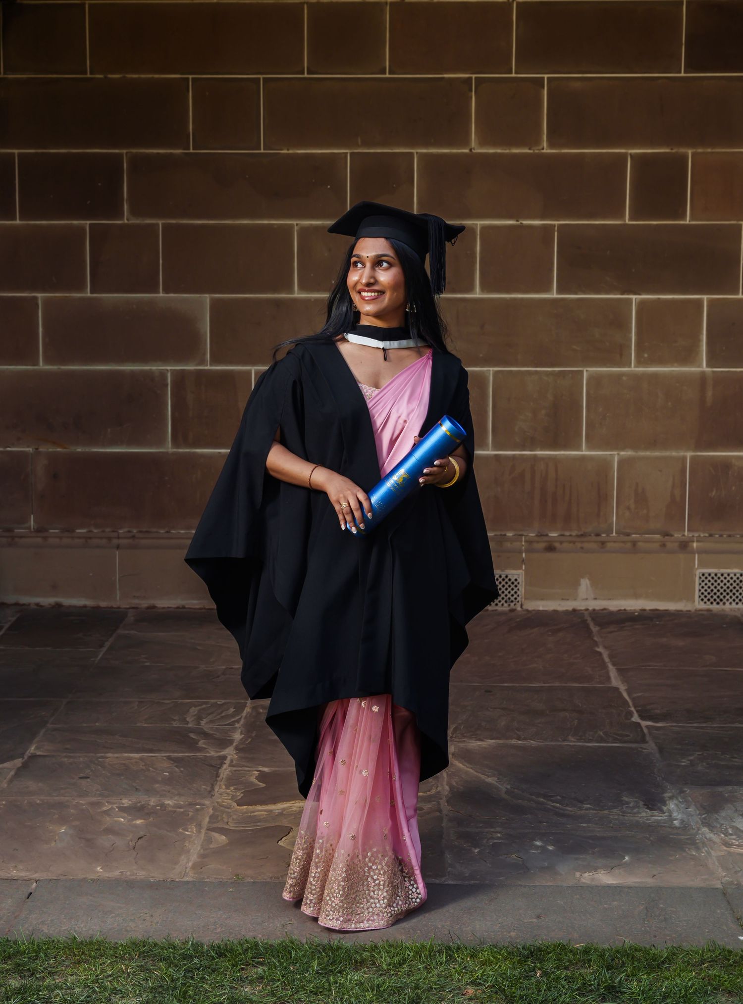 A graduate in black academic regalia poses against a brick wall in a series of celebratory graduation photos.