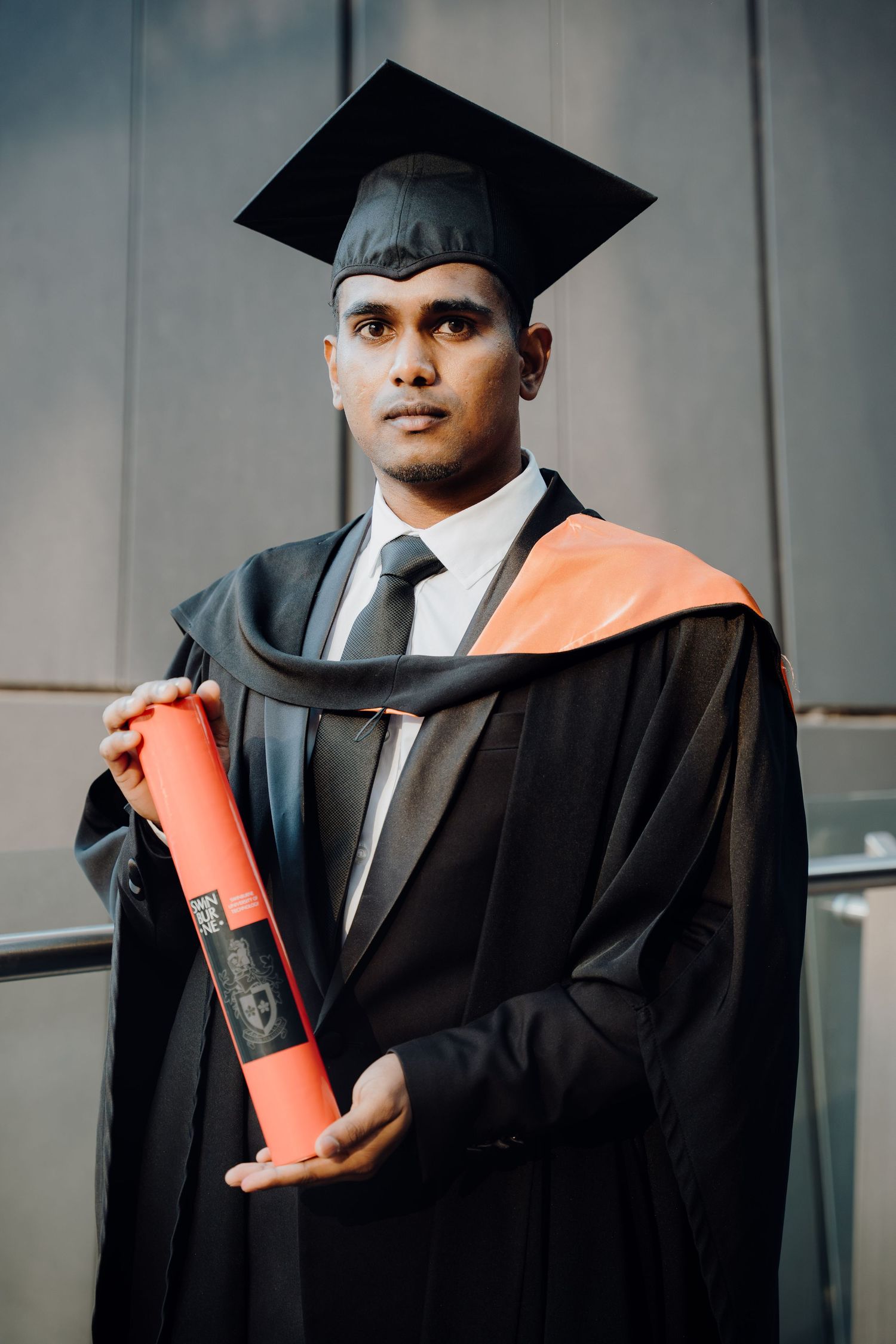 Young man in black graduation gown with orange trim and mortarboard holds diploma at university ceremony.
