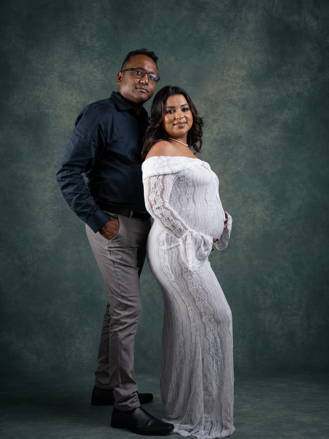 Couple posing for maternity photo shoot, woman in white lace gown cradling baby bump, man in dark shirt beside her.