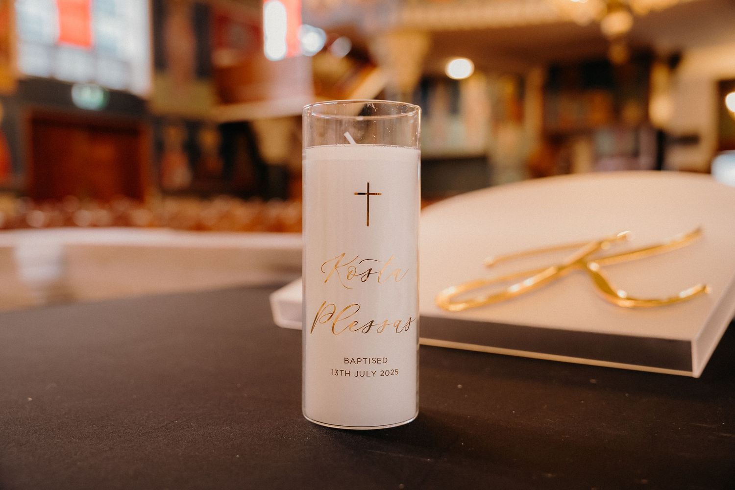 A white prayer candle with a cross symbol sits on a dark table with blurred warm lighting in the background.