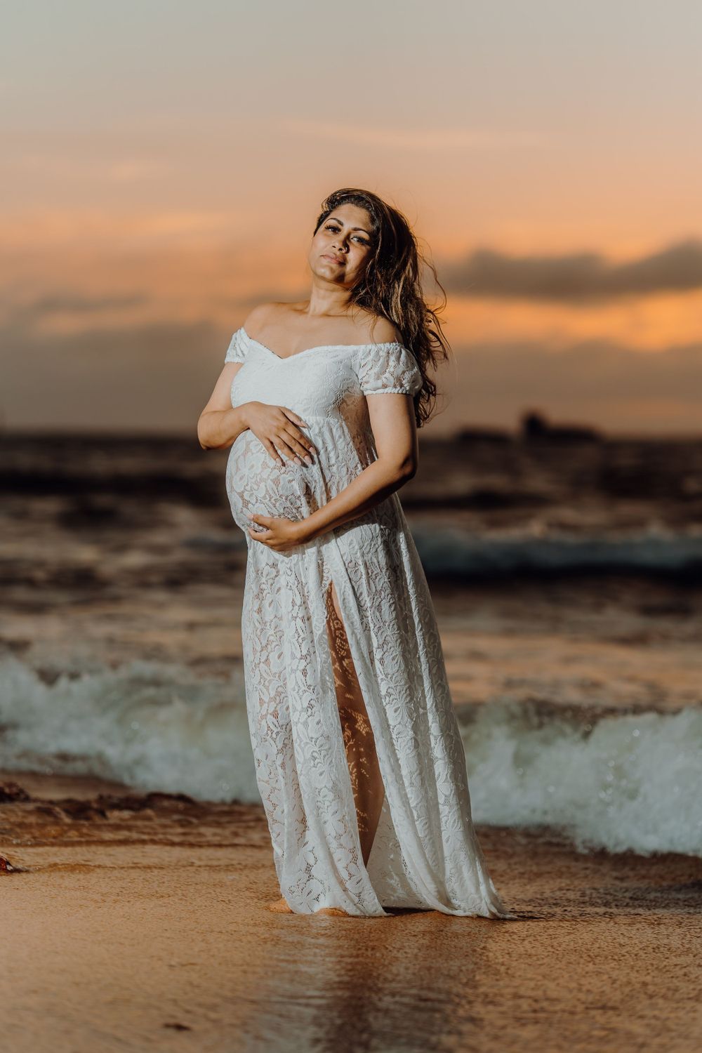 A person in a white lace dress poses on a beach at sunset with waves crashing in the background.