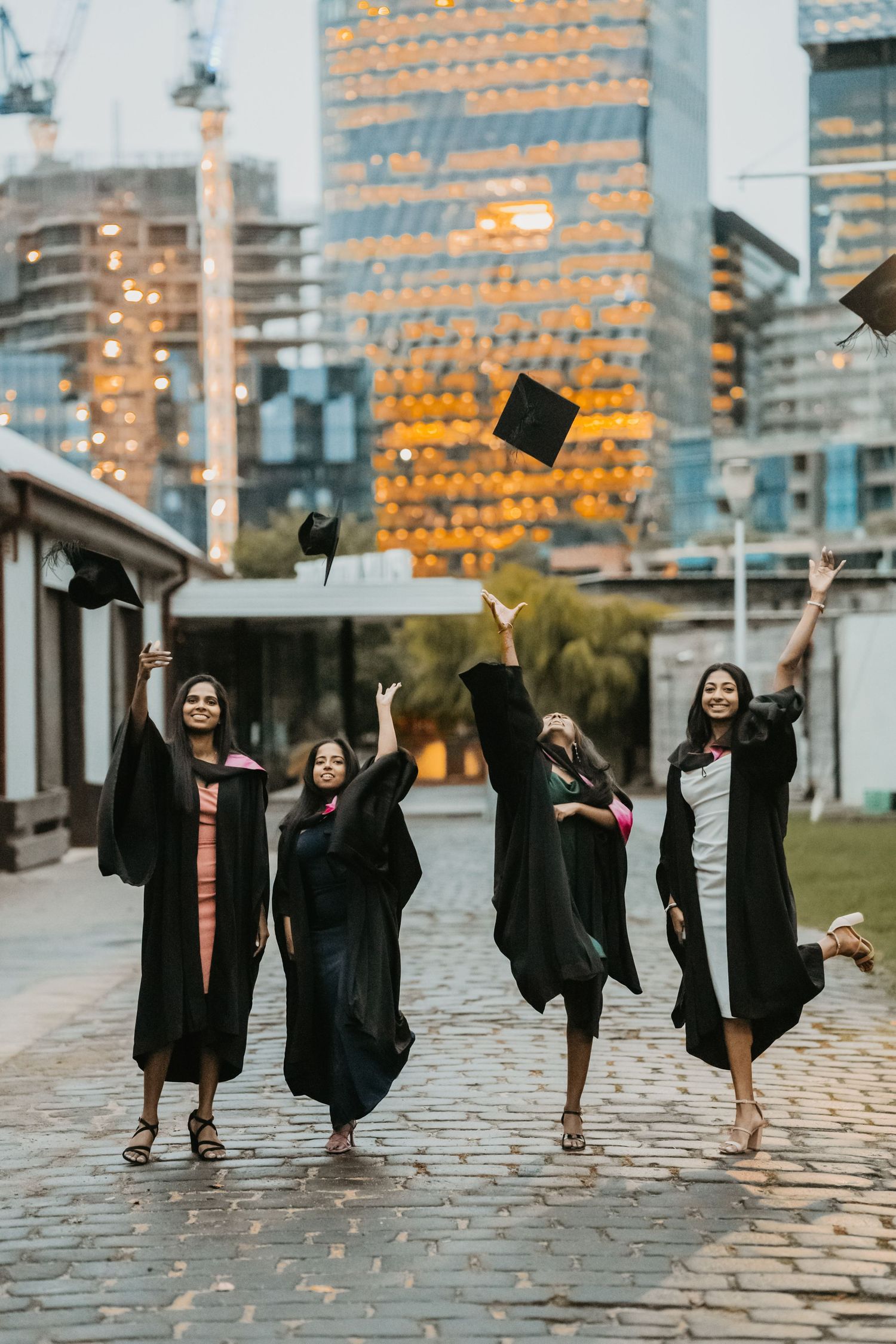 Two graduates in black gowns celebrate their graduation on a cobblestone path at dusk with city buildings in background.