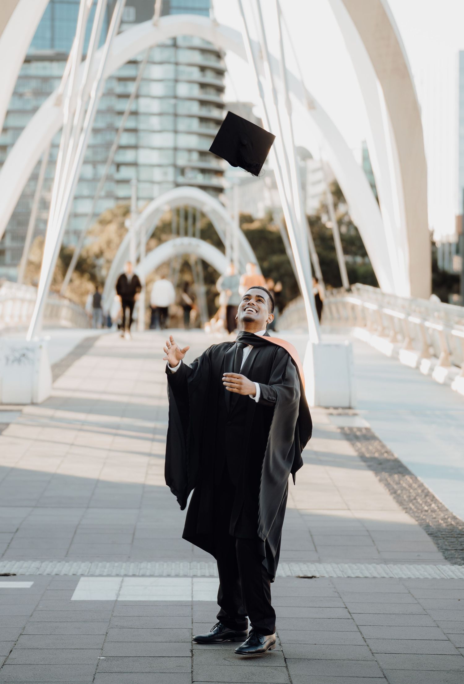 Graduate in black cap and gown poses on modern bridge with architectural white beams in background.