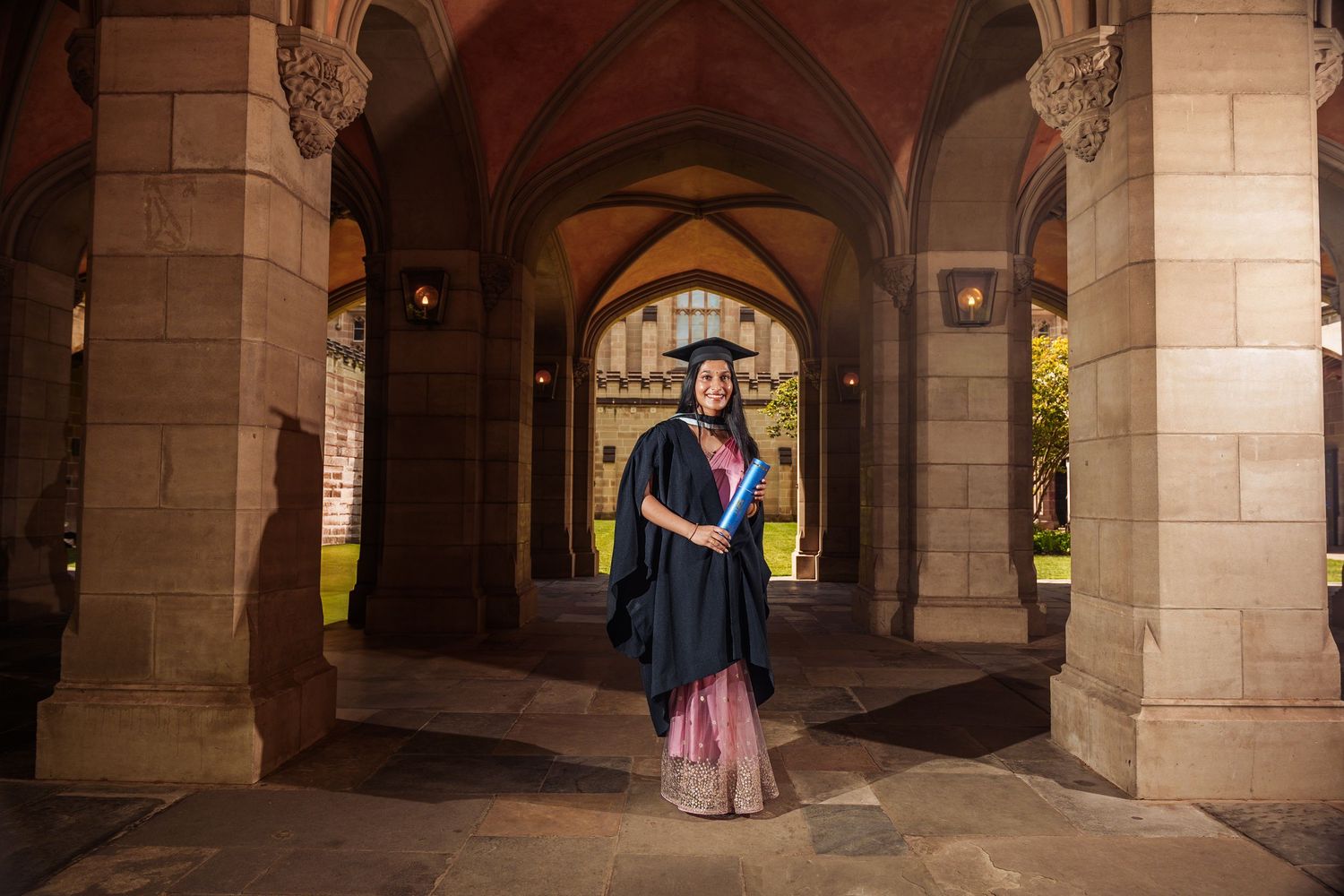 Graduate in cap, gown, and pink lehenga stands in gothic stone archway at university graduation.