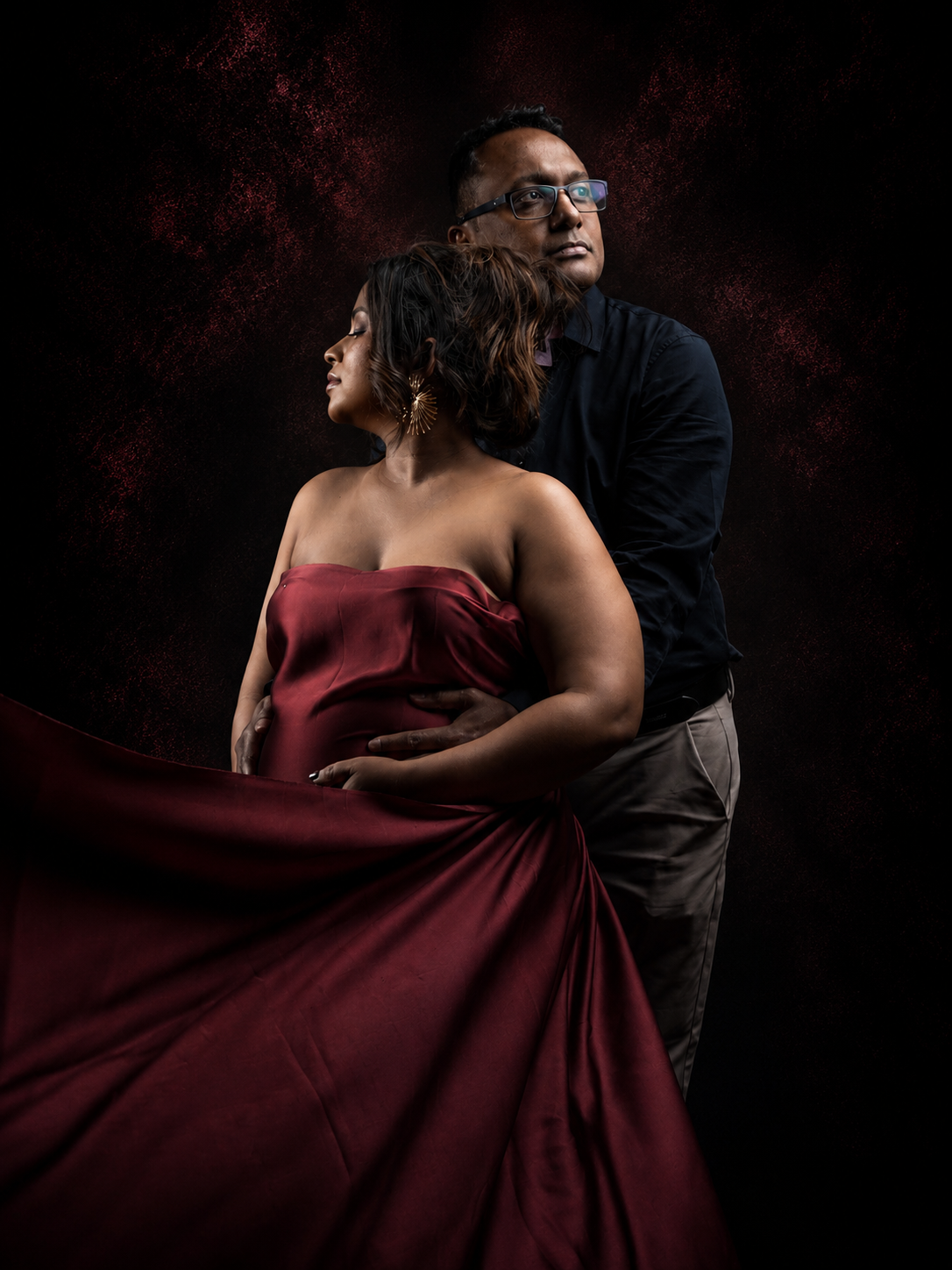Couple in dramatic studio portrait, woman in flowing red gown, man in black shirt, dark moody background.
