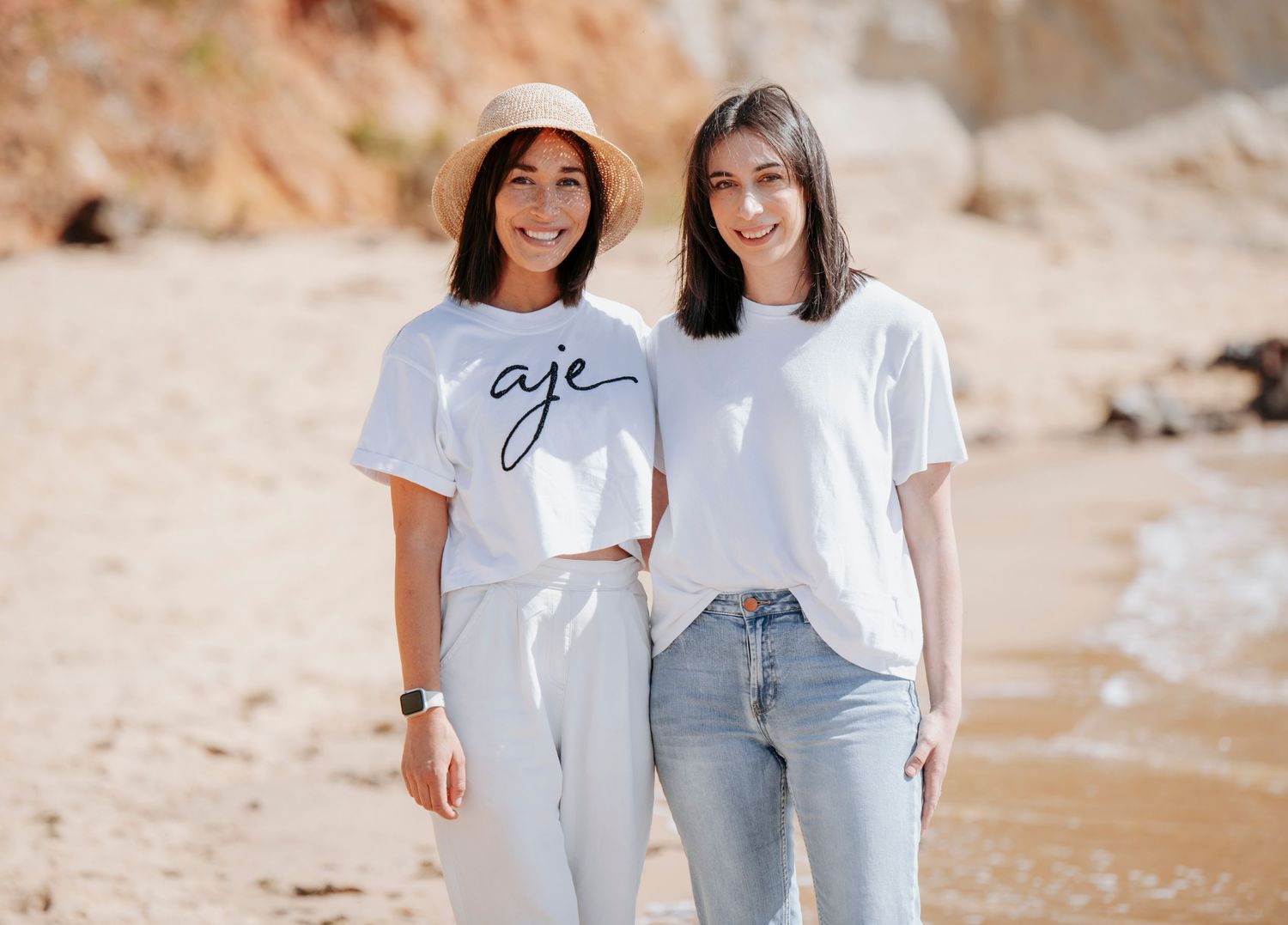 Two friends in casual summer outfits with white t-shirts and denim standing on a sandy beach.
