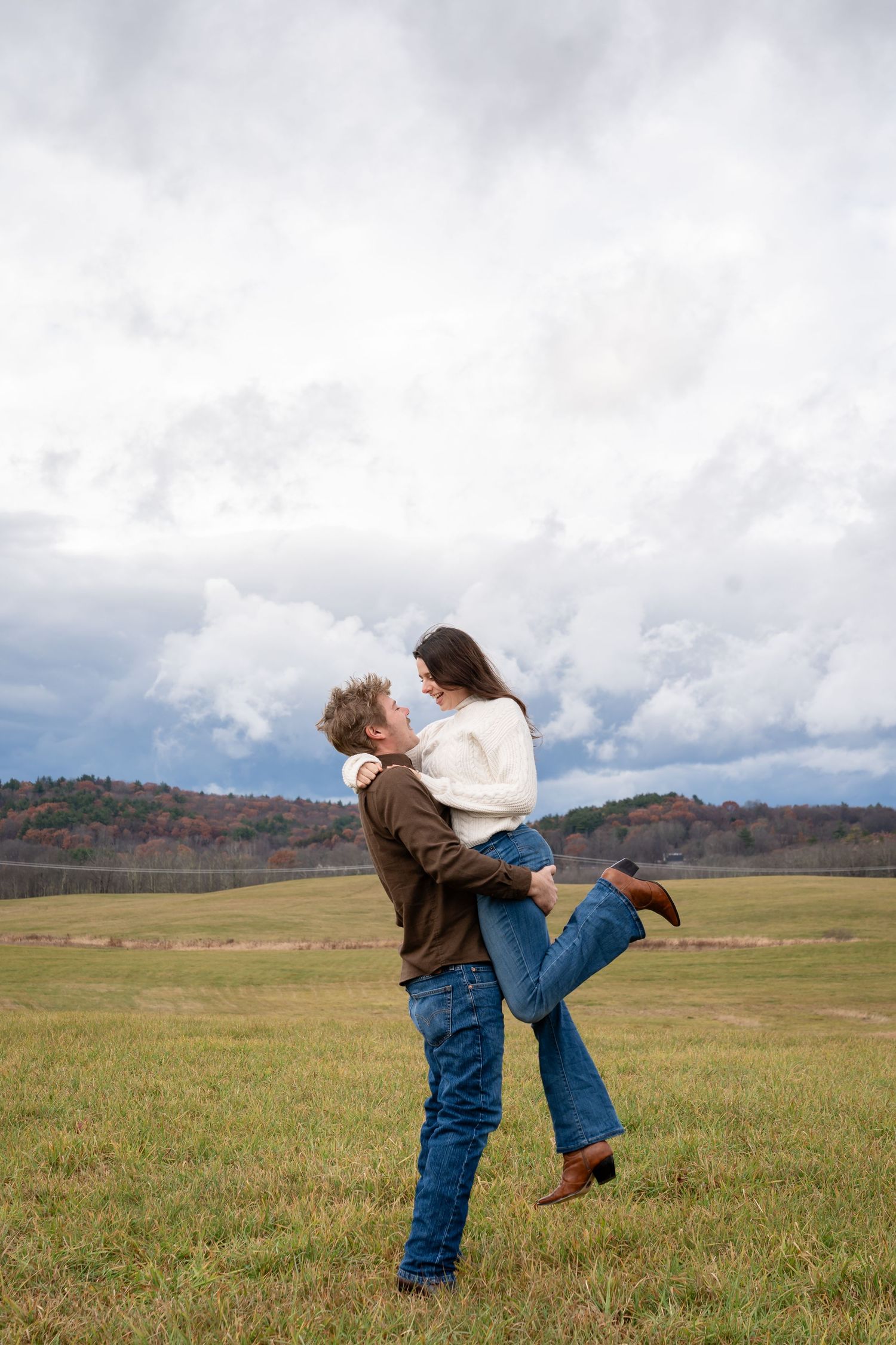 A man lifts a woman in an open field with dramatic cloudy skies and autumn hills in the background.