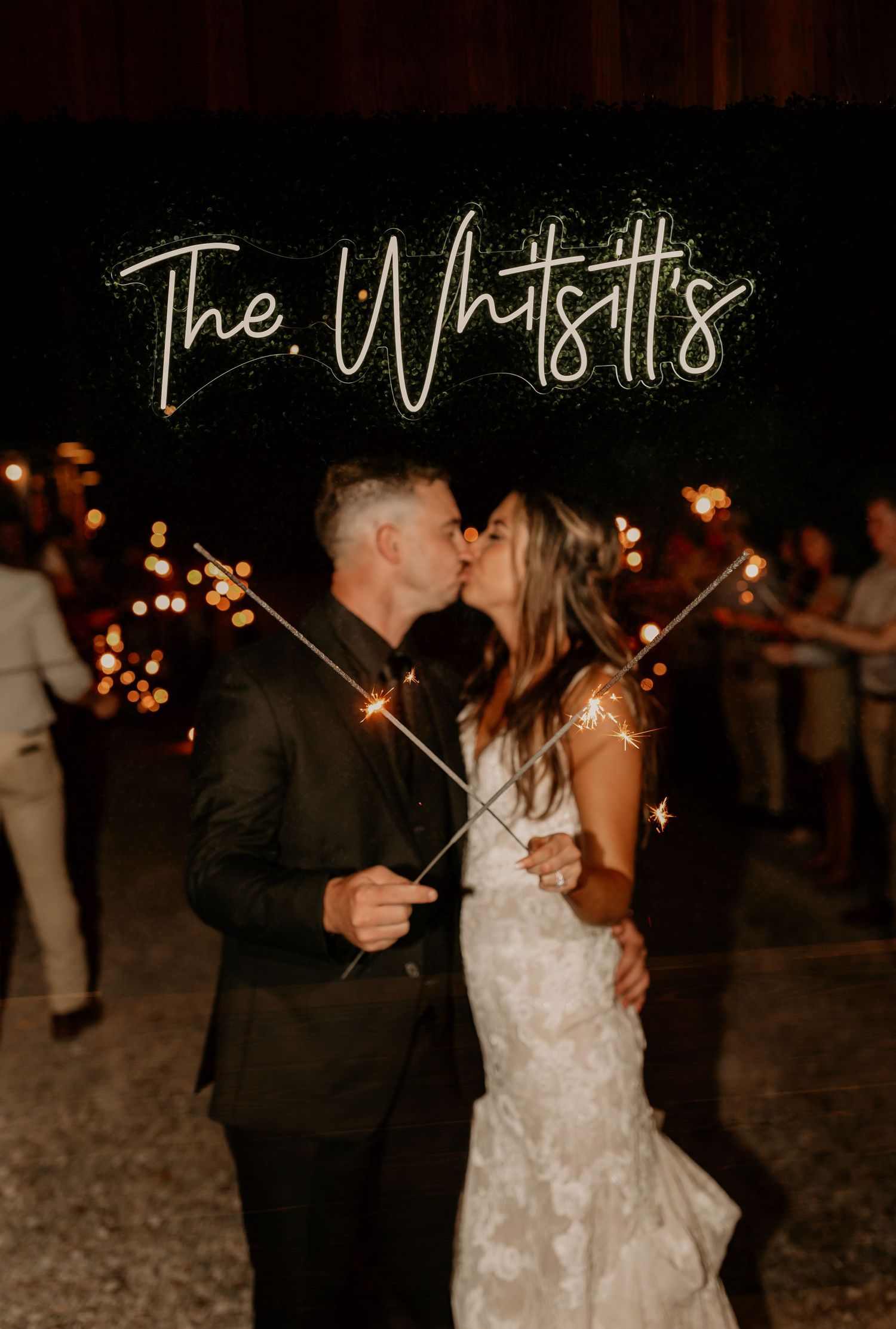 A couple shares a kiss under sparklers and a neon sign at their wedding reception.