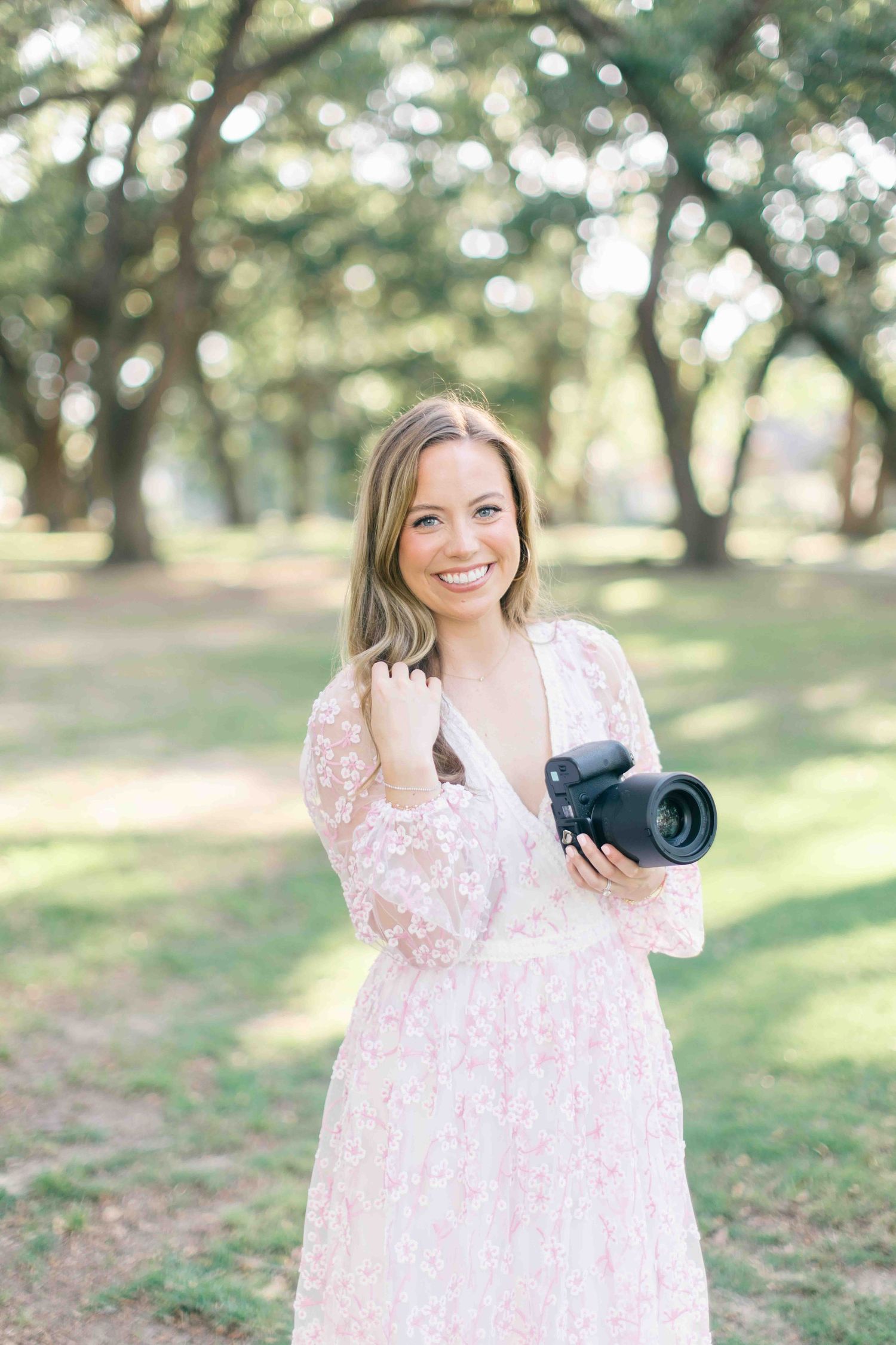 Alex Ishee, specializing in Fairhope photography in a pink floral dress poses outdoors with a professional camera in a sunlit park setting.