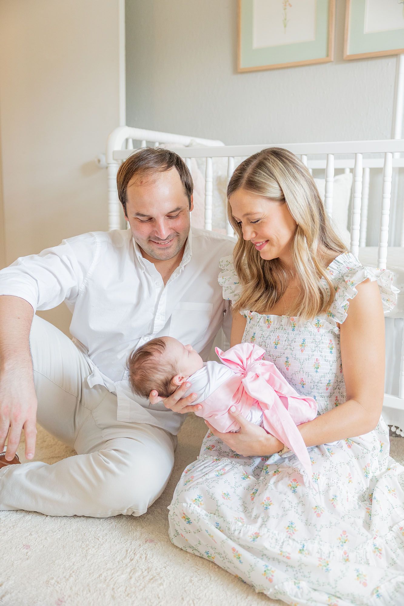 Mom and dad sitting in a nursery in front of the crib holding and staring at baby wrapped in a white swaddle with a pink bow.