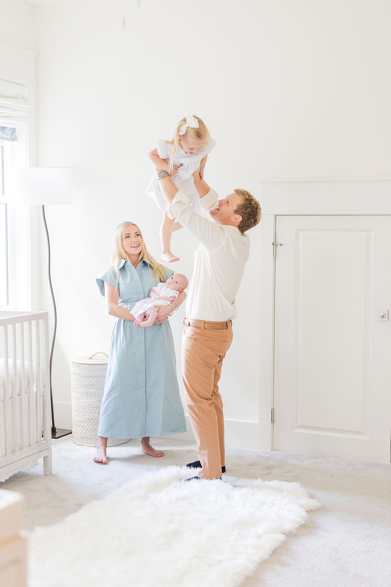 Dad playing airplane with his toddler daughter while mom holds baby in the background and smiles.