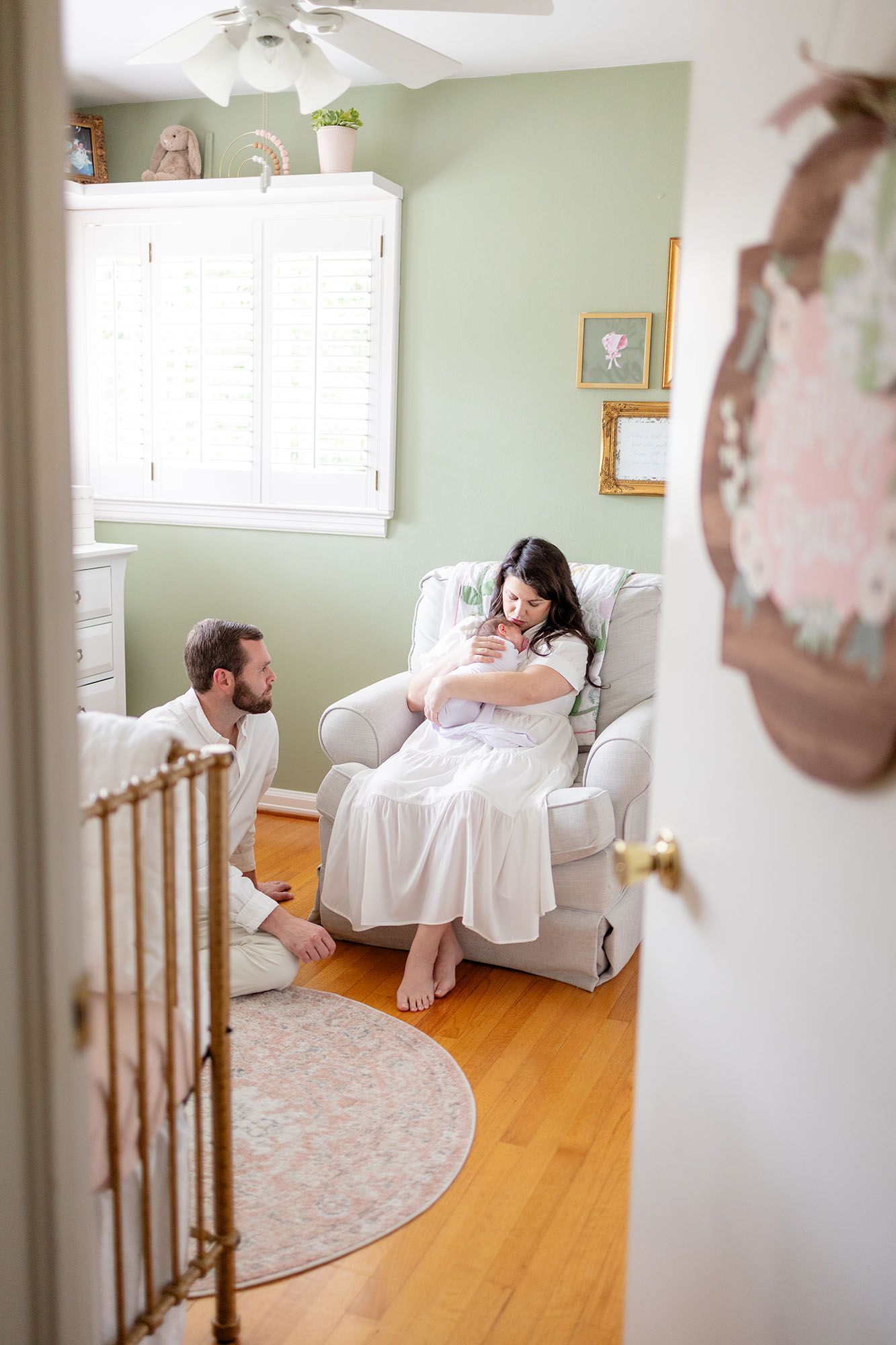 Photo by Alex Ishee Photography of mom rocking a baby in a green nursery while dad sits on the floor watching.