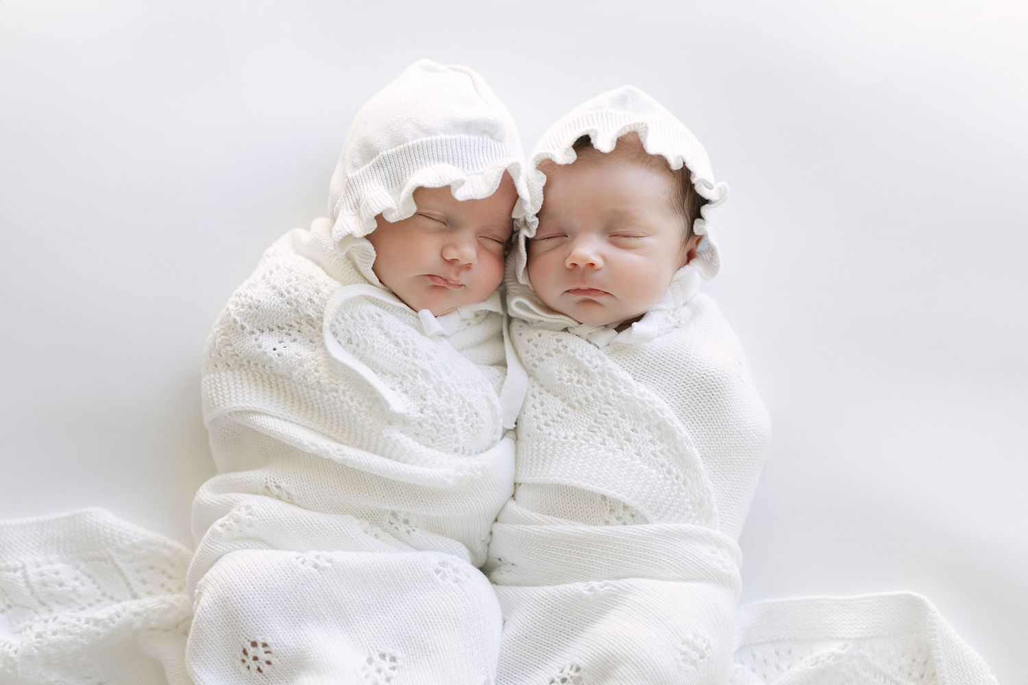 Twin baby girls wearing matching knit bonnets sleeping next to each other on a white sheet and swaddled in white blankets.
