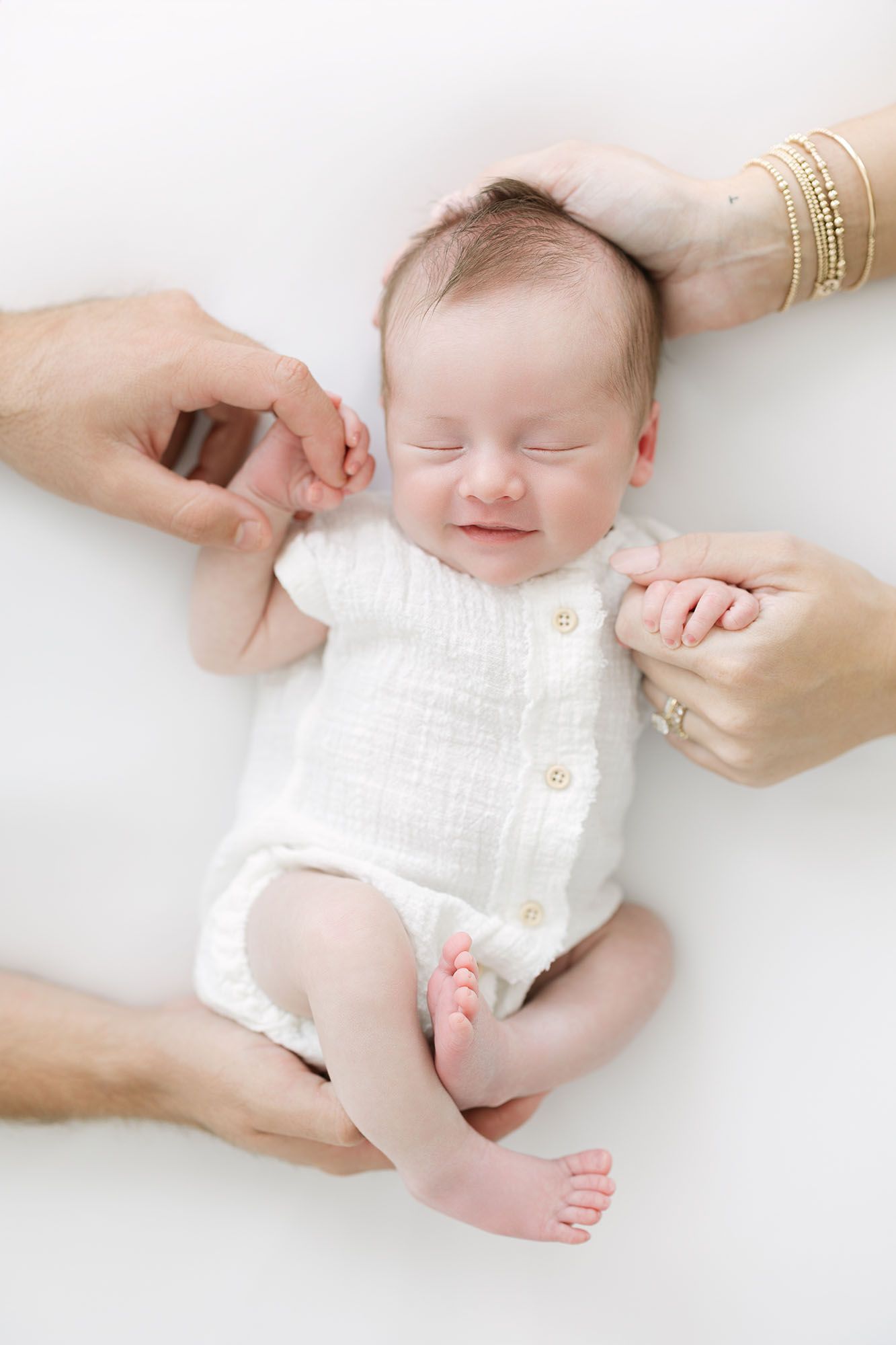 Baby smiling on white sheet and wearing a white linen button down onesie holding the fingers of his mom and dad.