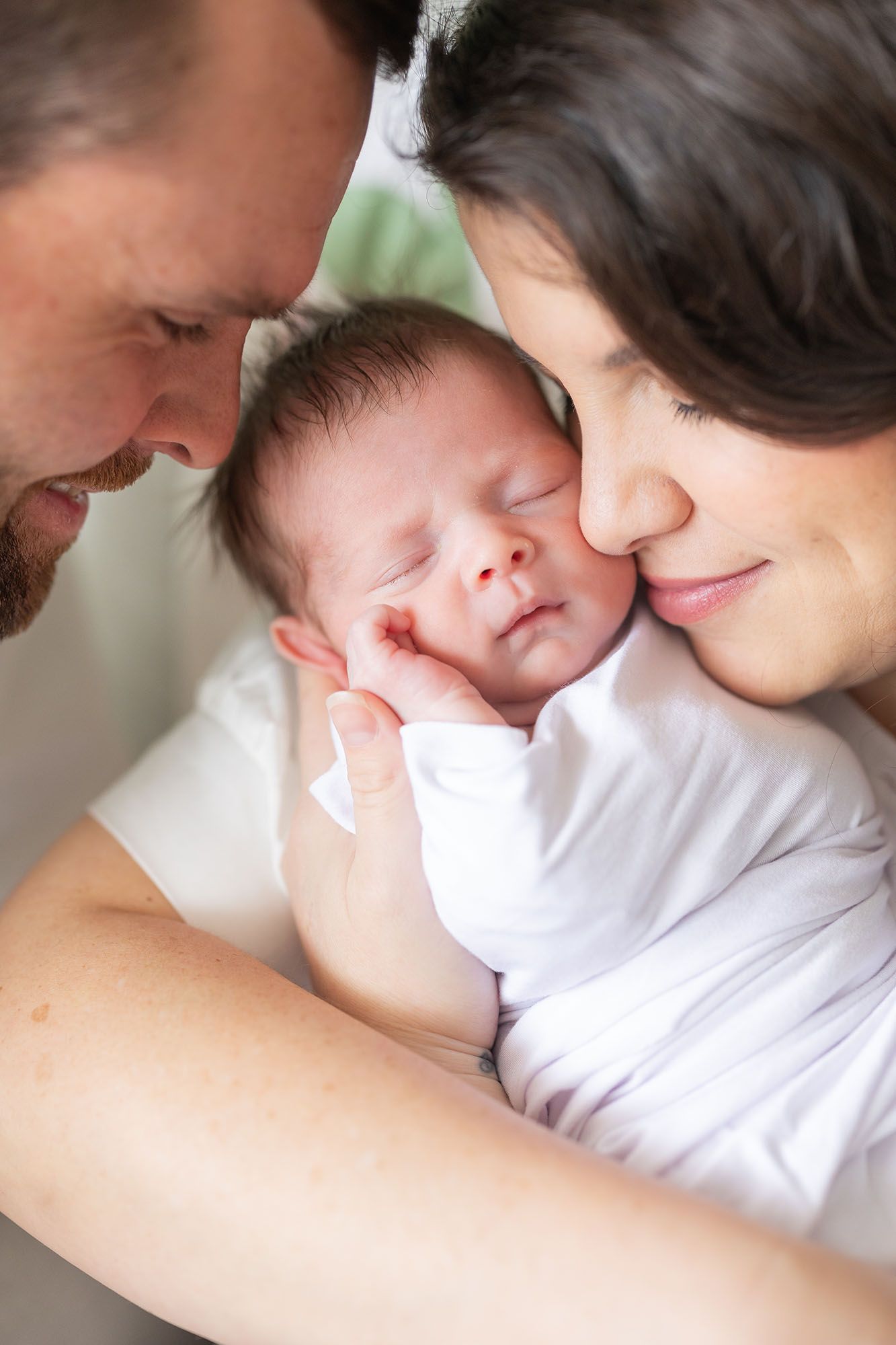 Mom and dad snuggling closely to baby girl wrapped in a white swaddle.